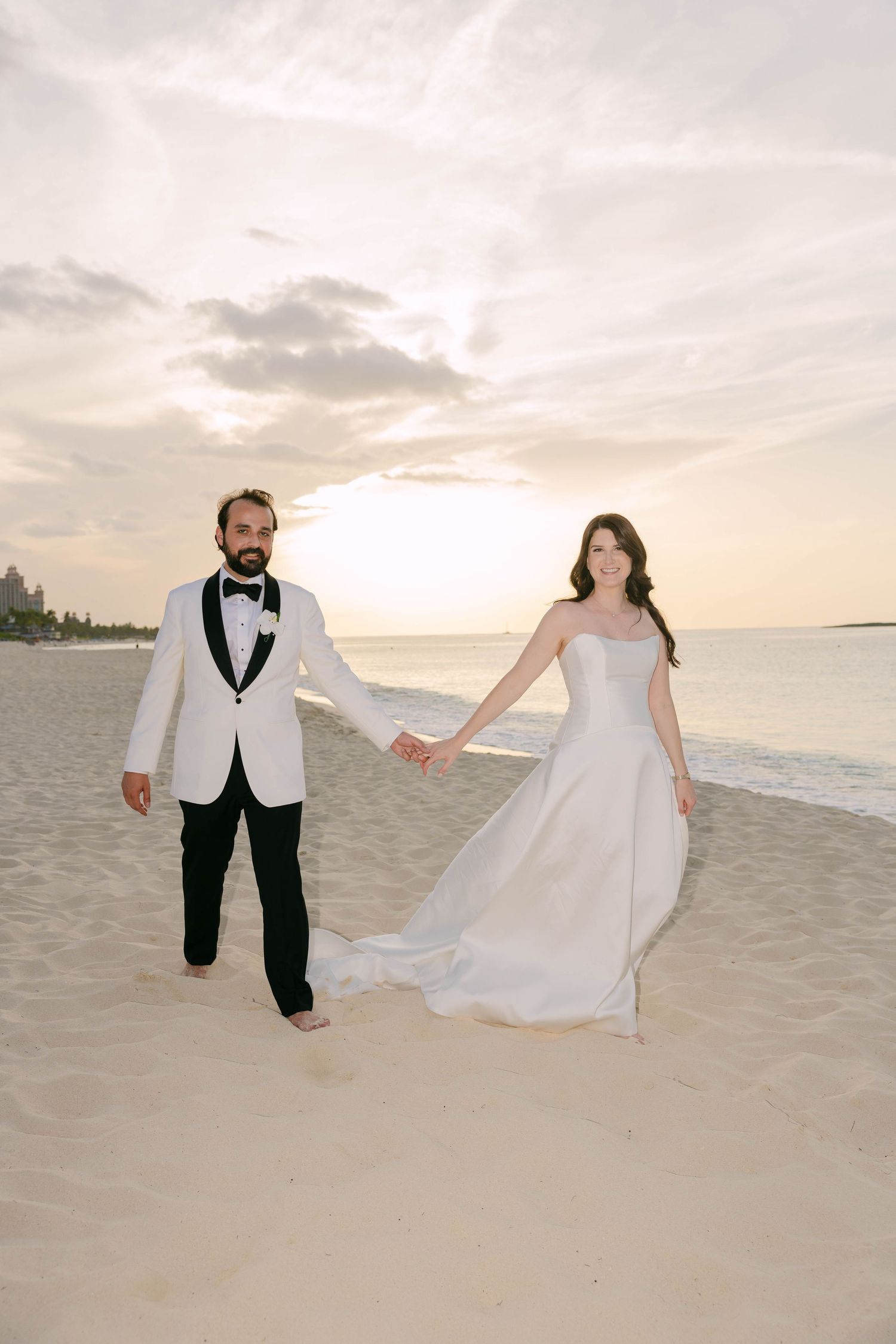 Bride and groom hold hands on a sandy beach at sunset, groom in white tuxedo jacket, bride in elegant strapless gown.