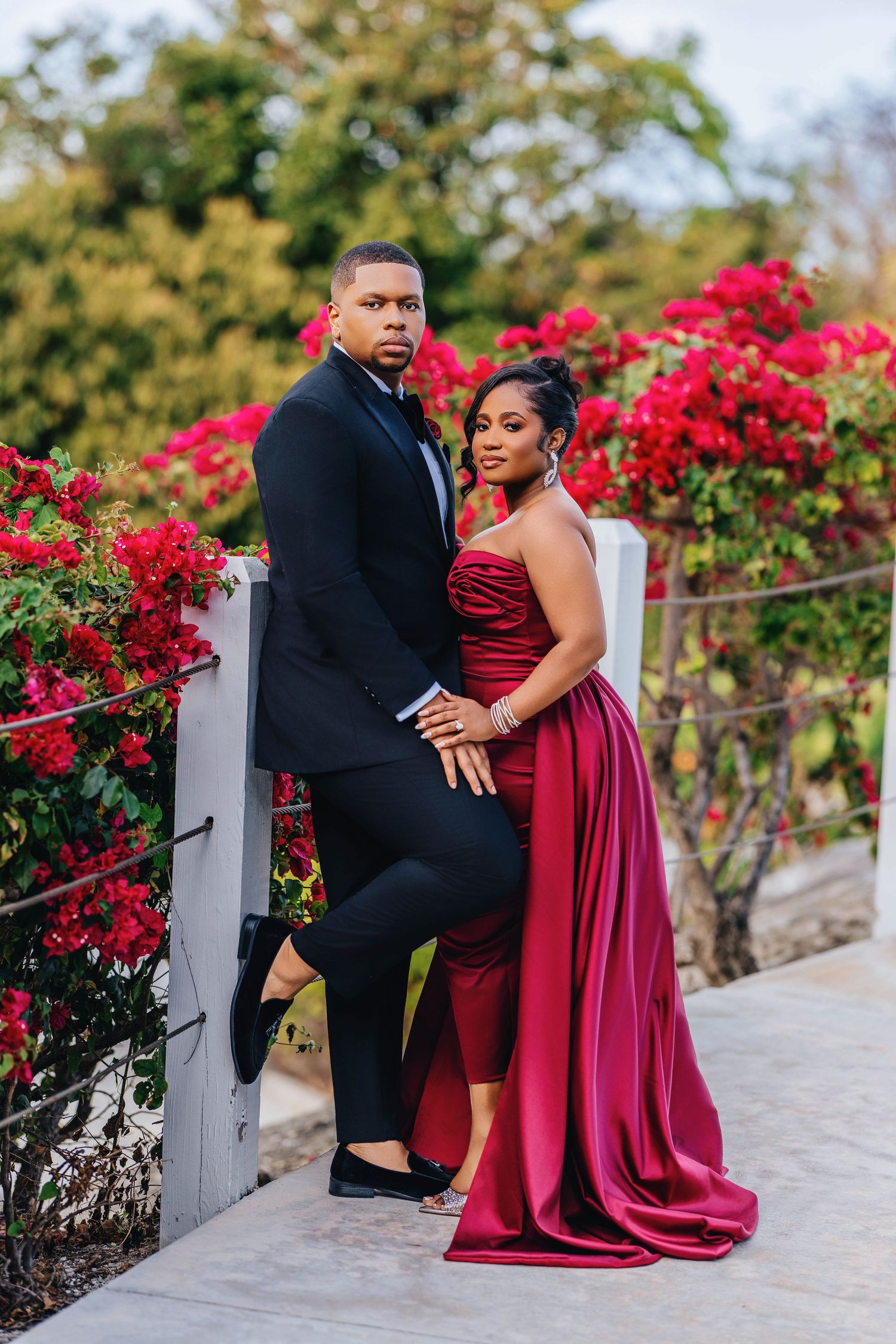 Formal couple posing by red bougainvillea flowers on outdoor pathway.