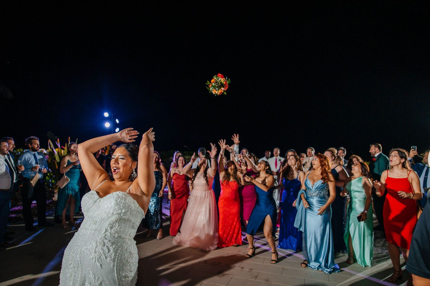 Bride tosses colorful bouquet to excited wedding guests gathered outdoors at night under bright lights.