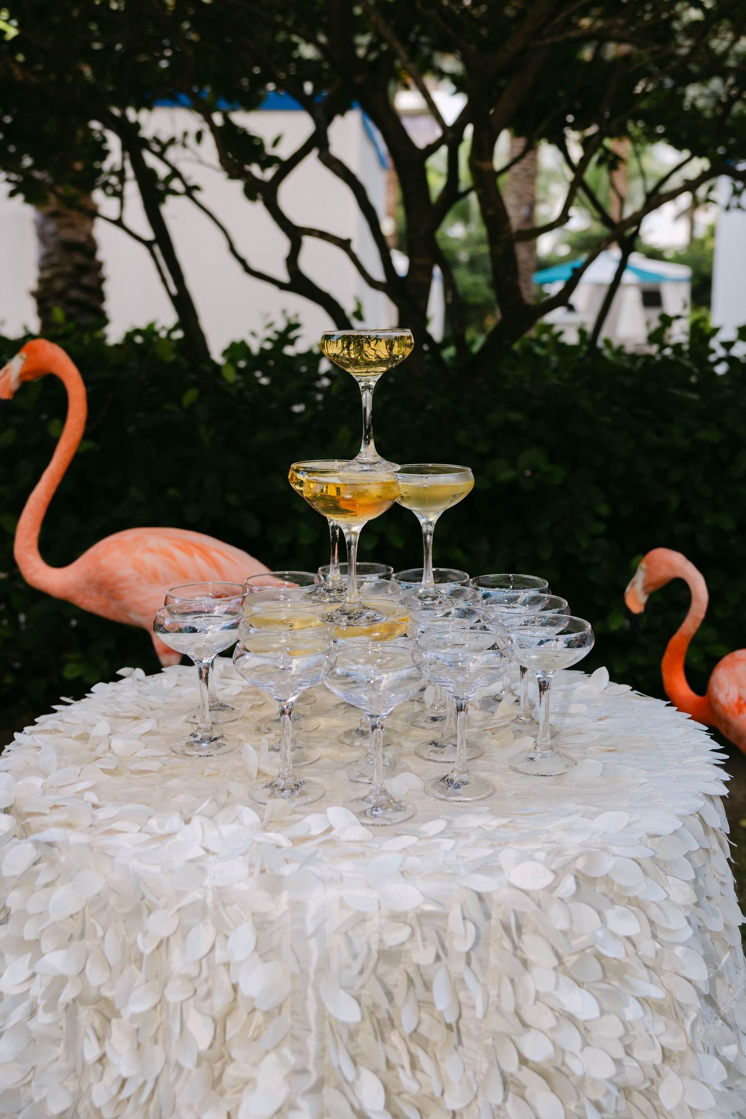 Champagne tower on white feather tablecloth flanked by pink flamingo decorations at an outdoor wedding reception.