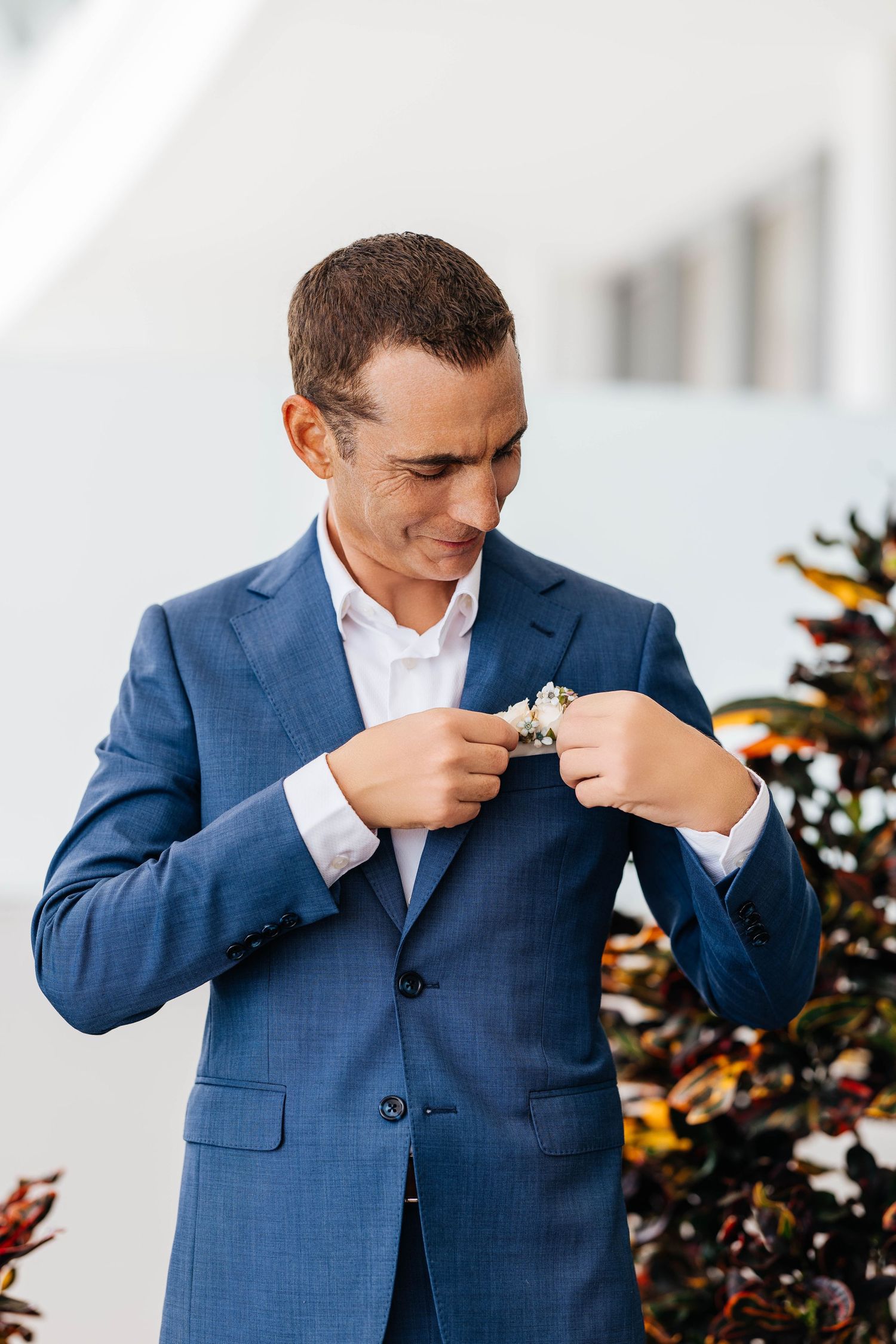 A groom in a blue suit pins a boutonniere to his lapel, smiling outdoors near colorful foliage.