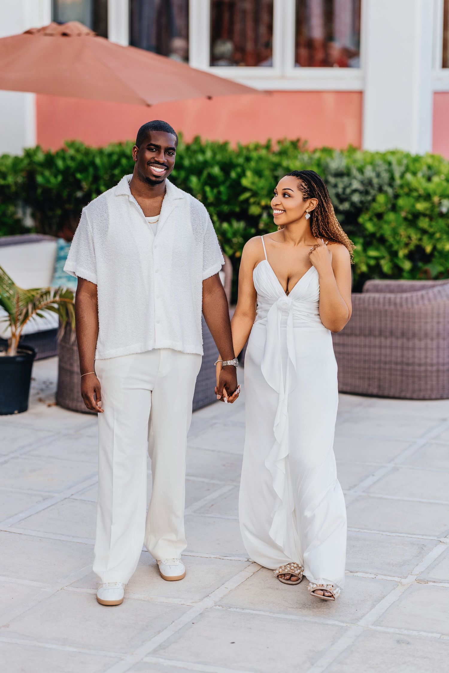 A happy couple in coordinating white outfits walk together on a tiled patio near a pink building.