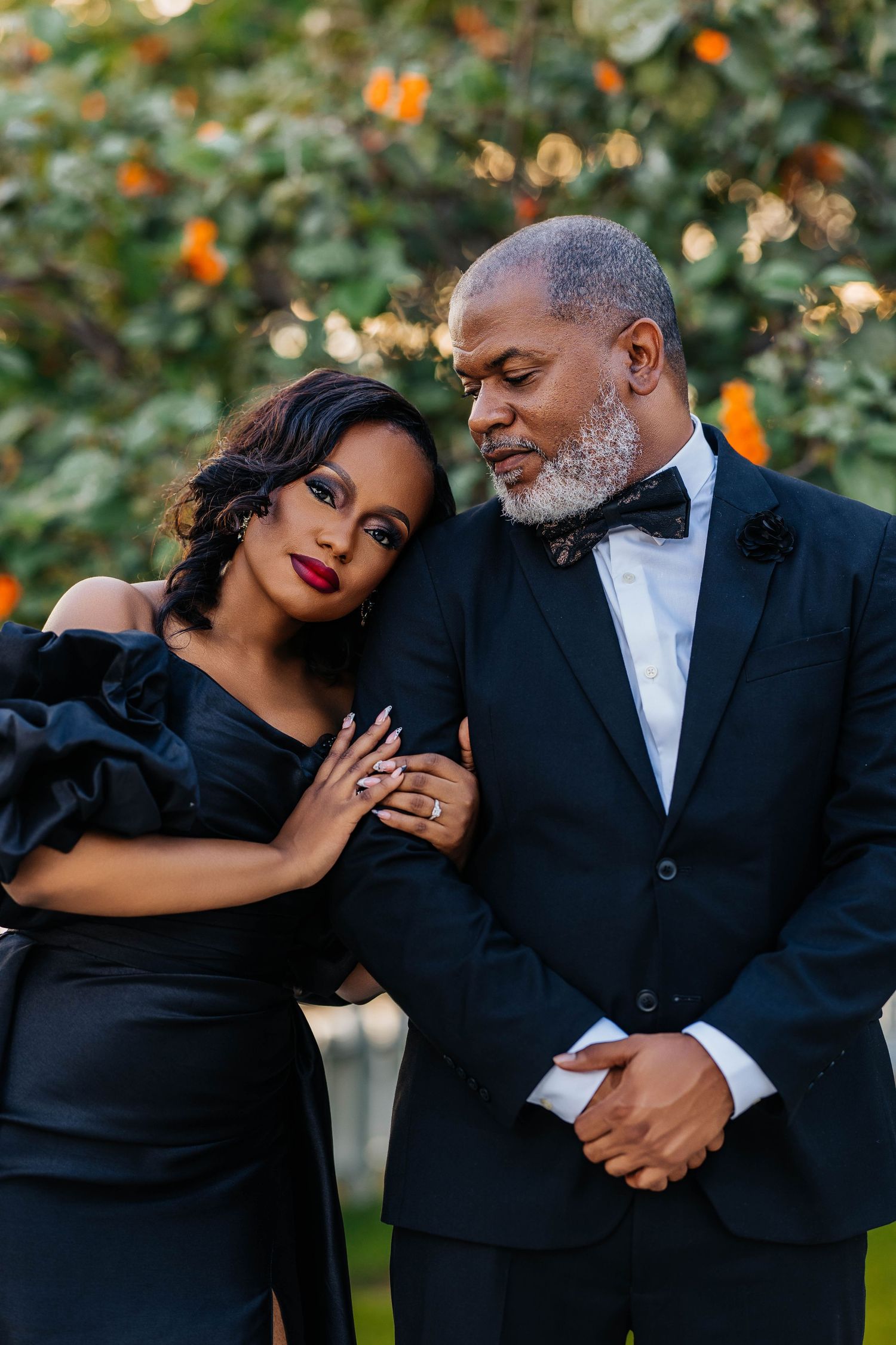 A romantic outdoor photo series showing a couple in formal black attire embracing against a backdrop with orange flowers.