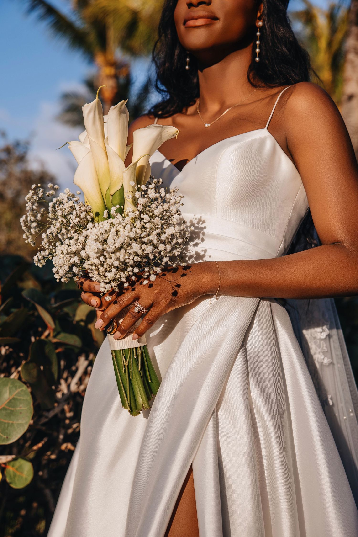 Bride in white satin gown holding calla lily and baby's breath bouquet outdoors with palm trees.