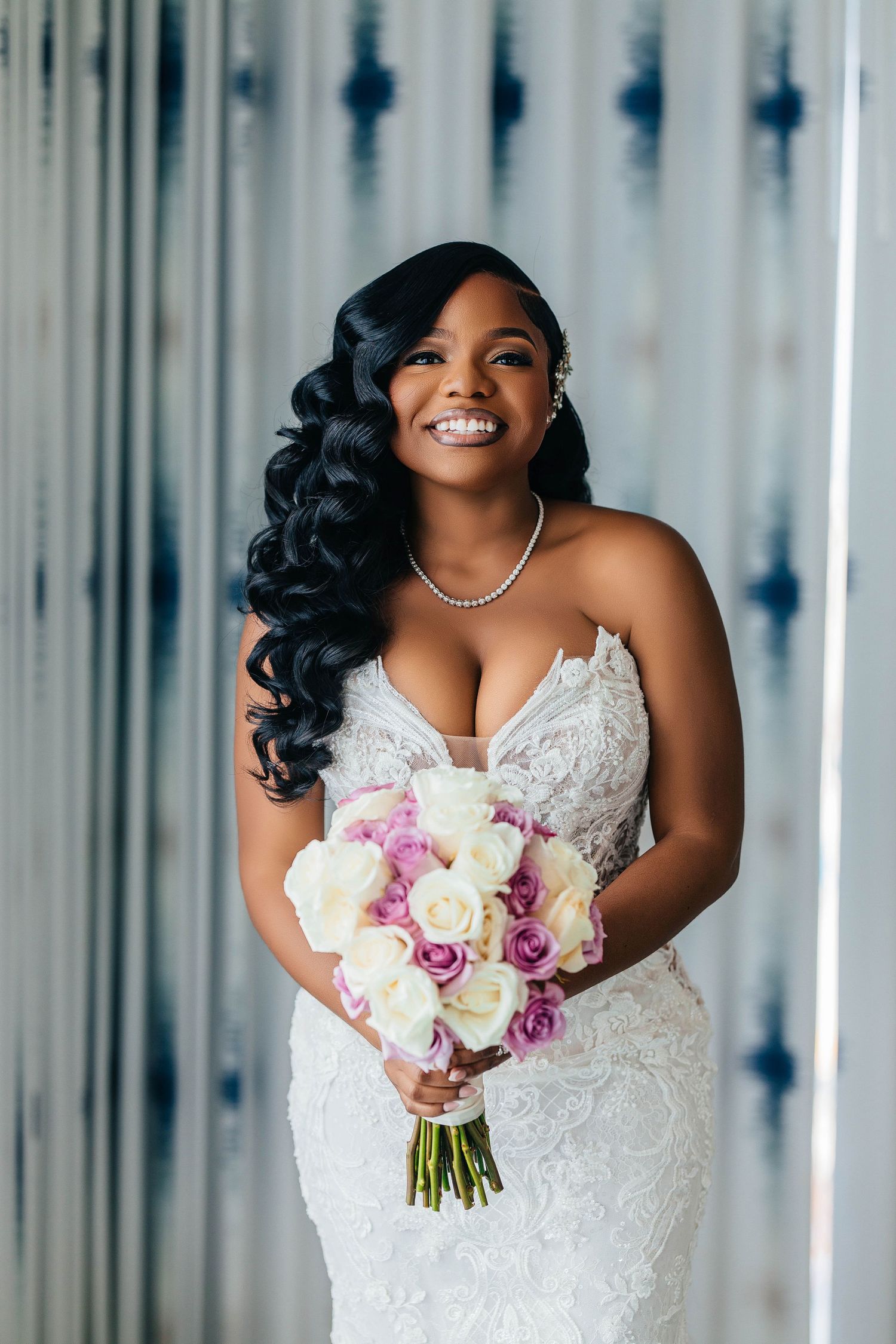 Smiling Black bride in lace mermaid gown holding white and purple rose bouquet with pearl necklace.