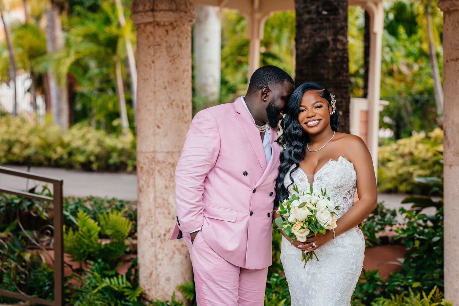 Wedding couple shares intimate moment in pink and white attire under garden colonnade.