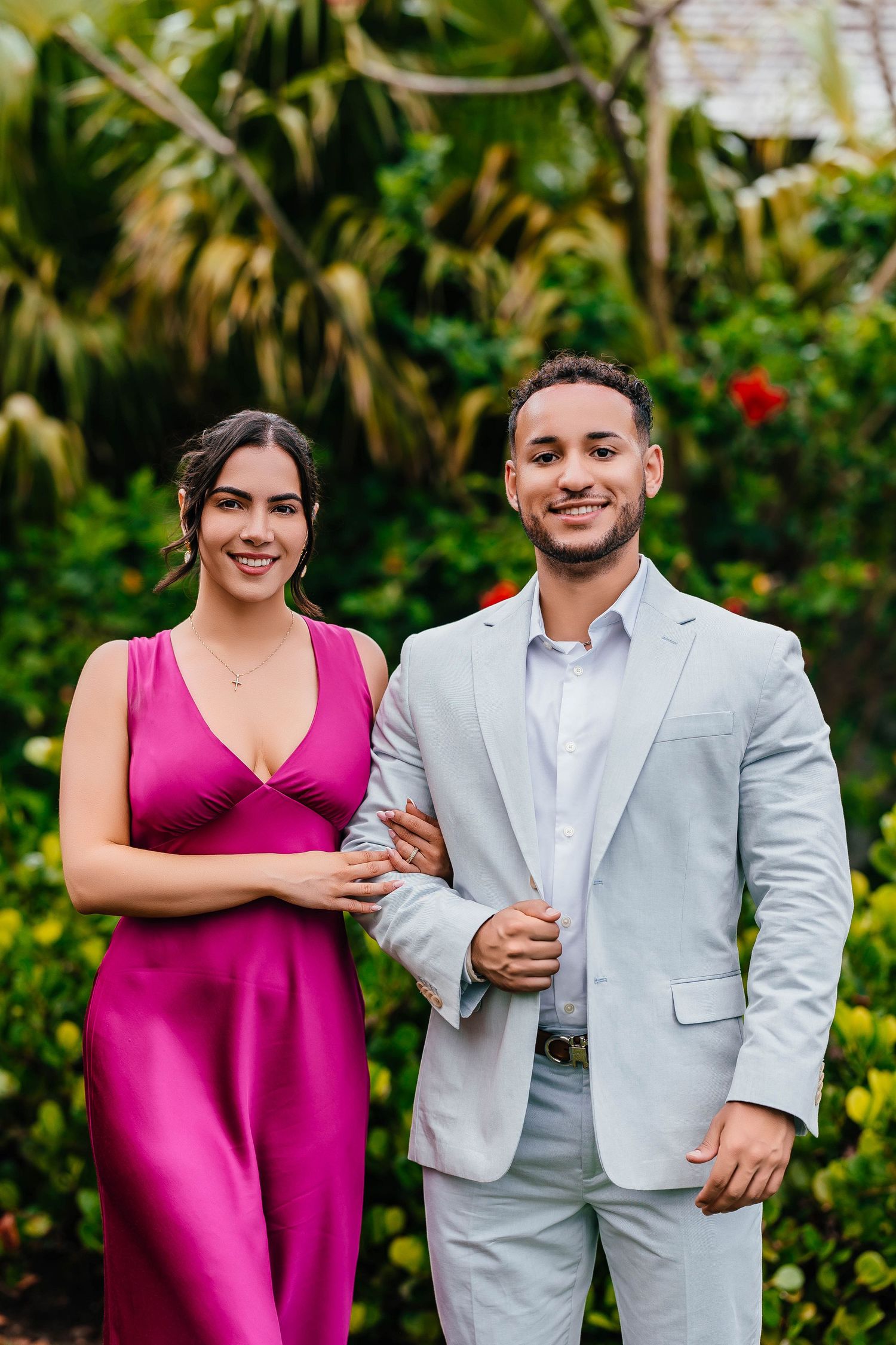 A couple in pink and gray formal wear poses together in a tropical garden setting.
