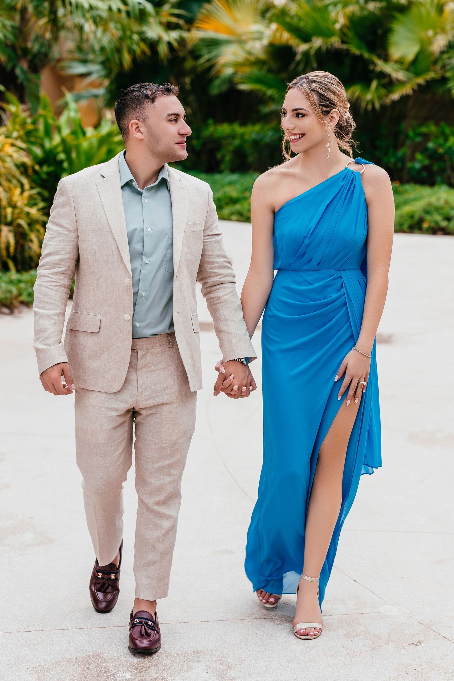Stylish pair walks hand in hand, wearing coordinated formal attire against lush tropical backdrop.
