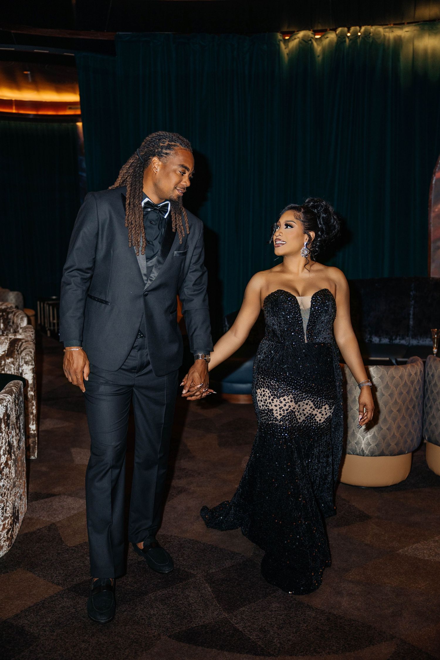 Couple in elegant black formal wear poses together against a dark teal backdrop.