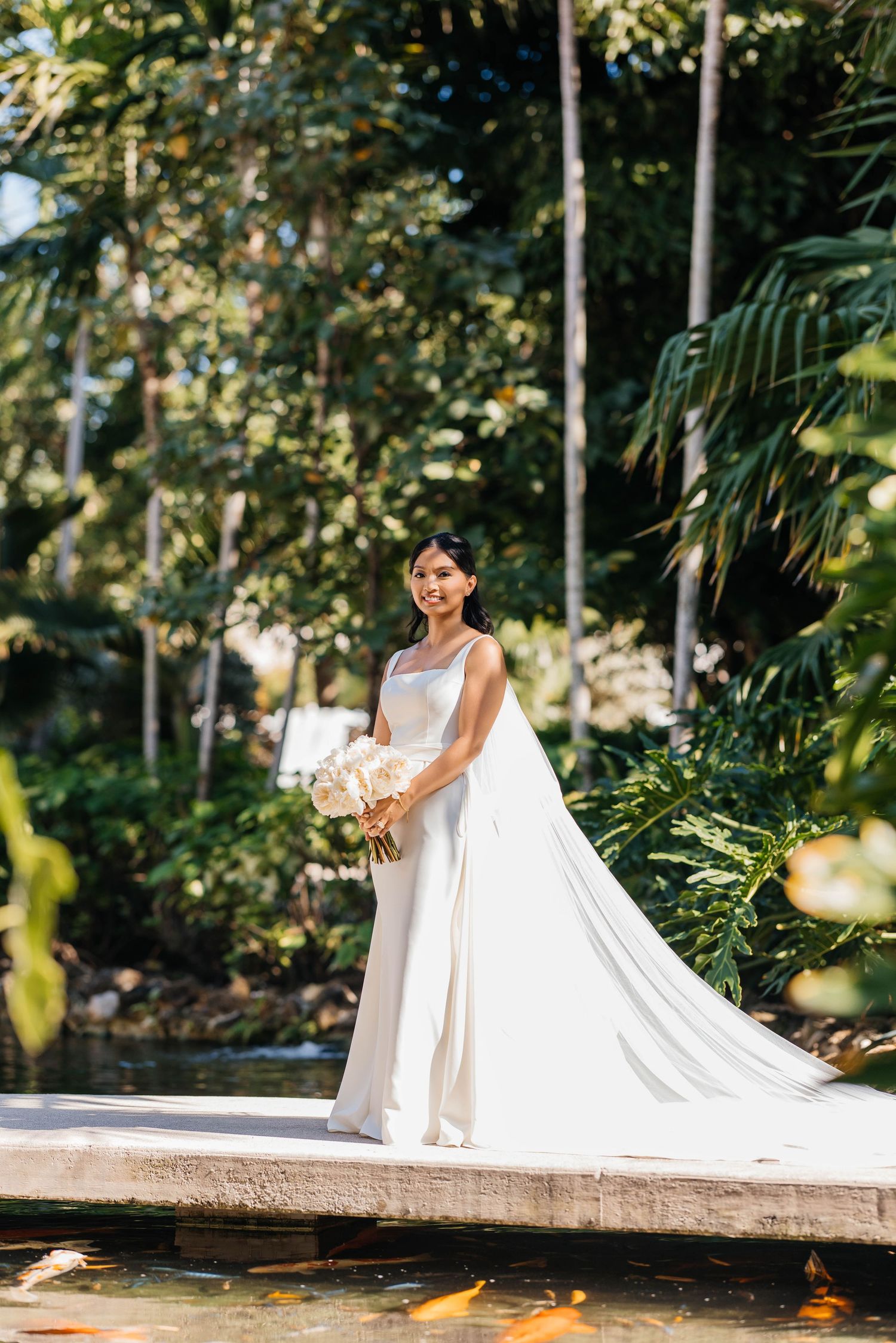 A bride in an elegant white wedding gown with a flowing cape stands in a lush tropical garden setting near a pond.