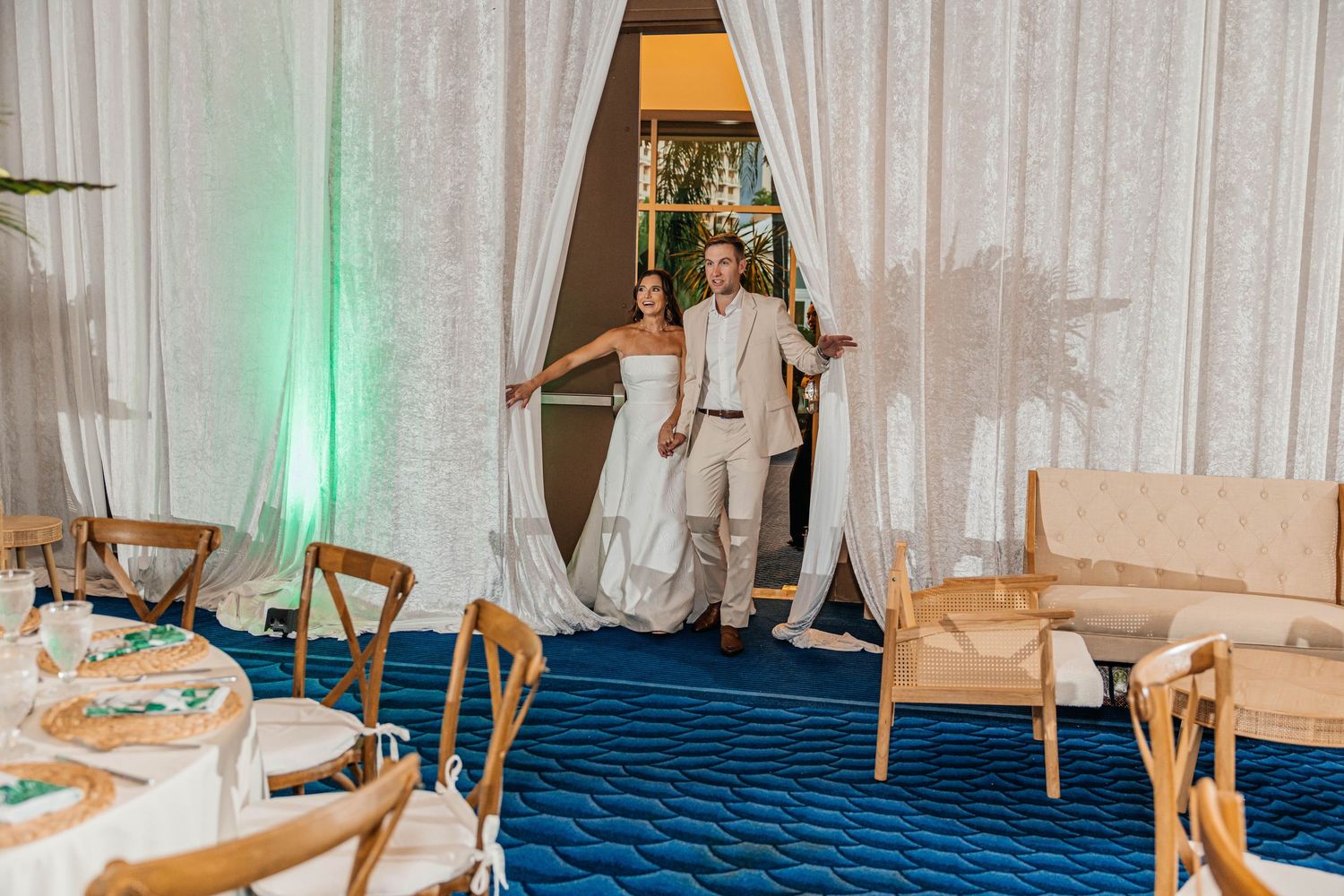Bride and groom make grand entrance through white draped doorway at elegant blue-carpeted wedding reception venue.