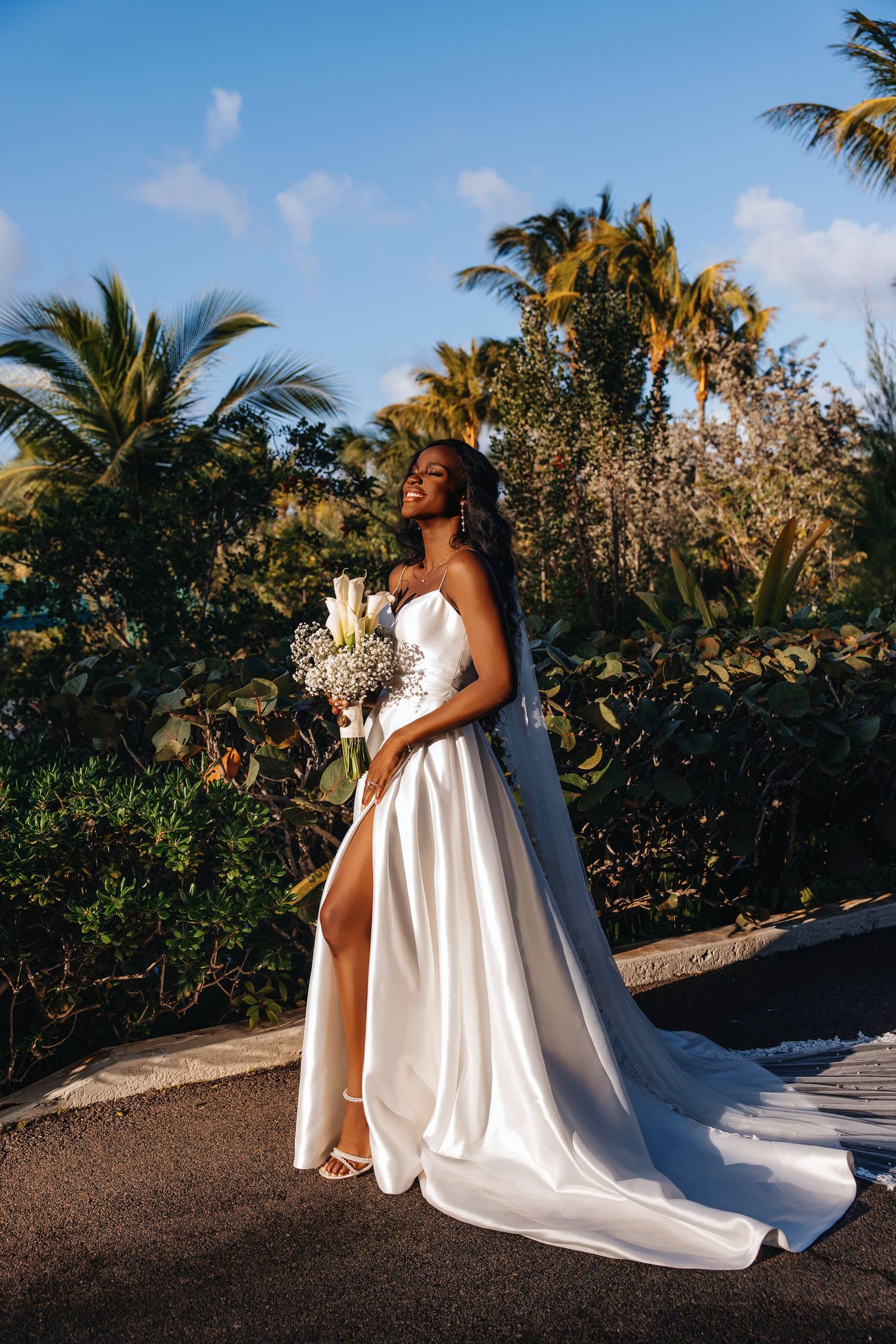Bride in white satin gown with slit holds bouquet outdoors, palm trees and blue sky in background.