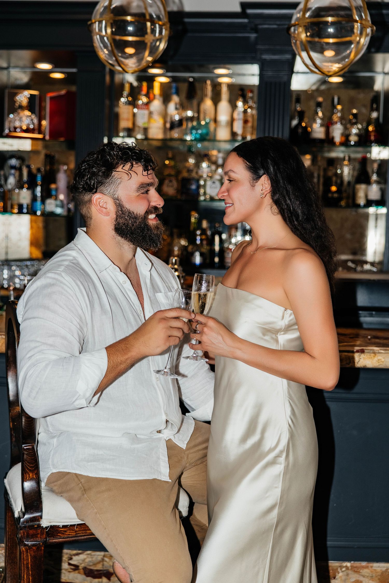 A couple shares a toast at an elegant bar with golden pendant lights.