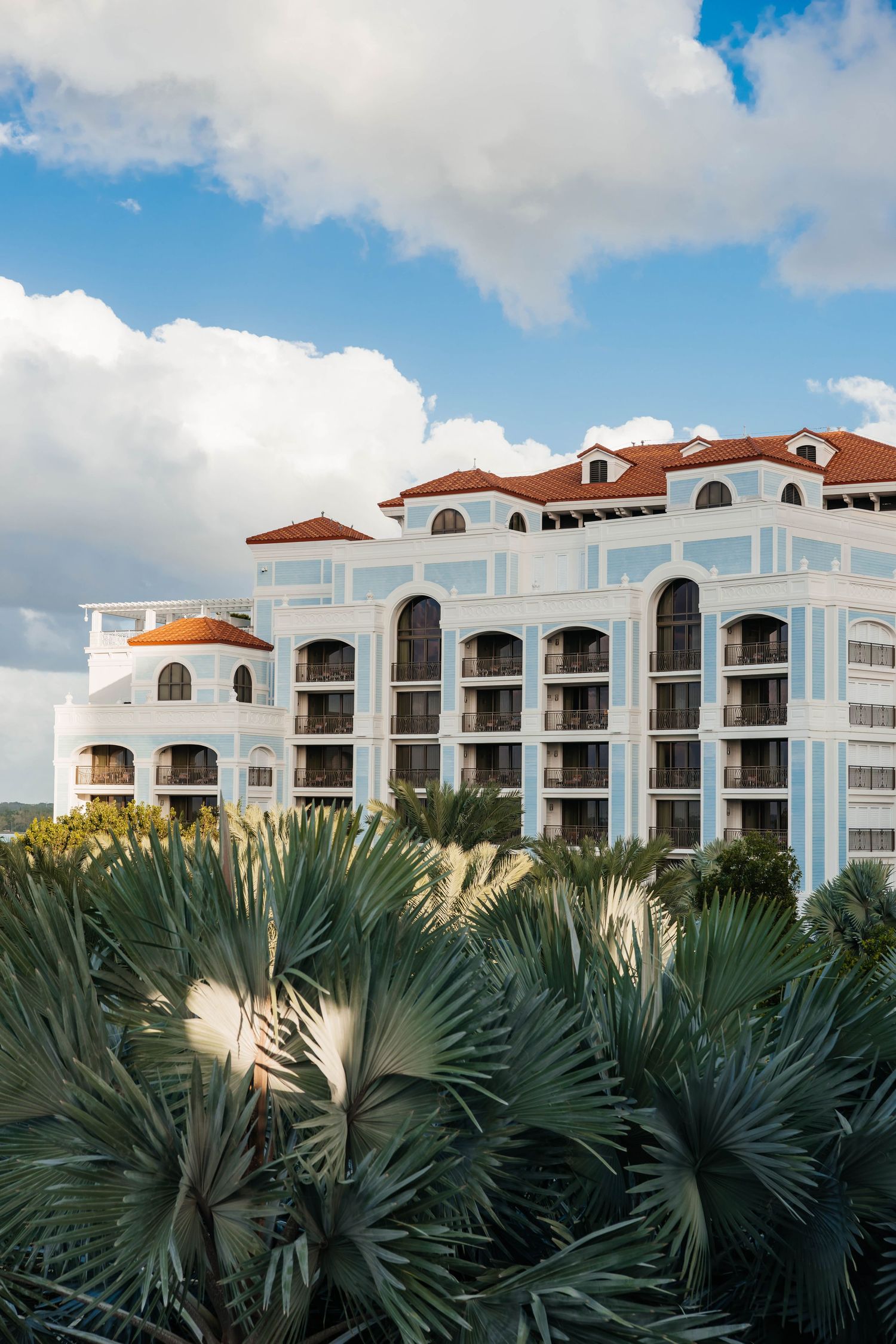 White Mediterranean-style resort with red tile roofs and balconies surrounded by lush palm trees under a blue sky.