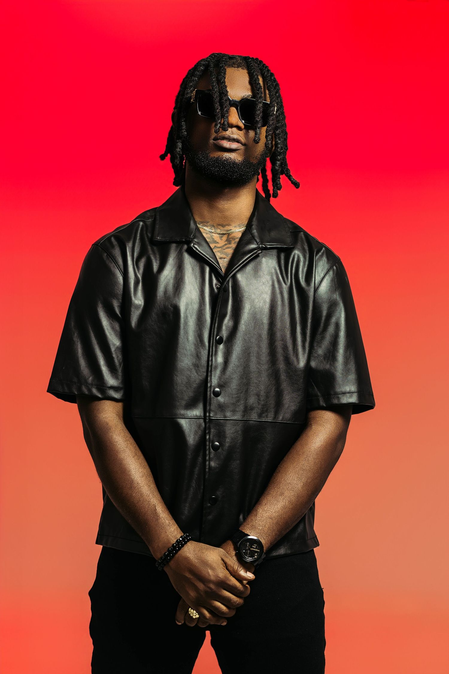 Artist wearing black leather shirt and sunglasses poses against vibrant red background.