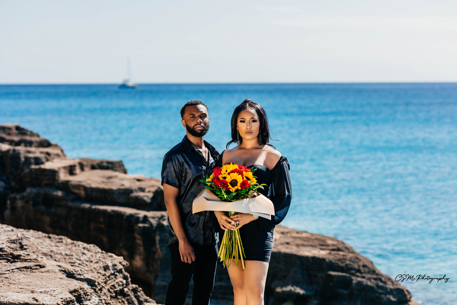 Couple posing together on rocky ocean cliffs with vibrant sunflowers and blue water backdrop.
