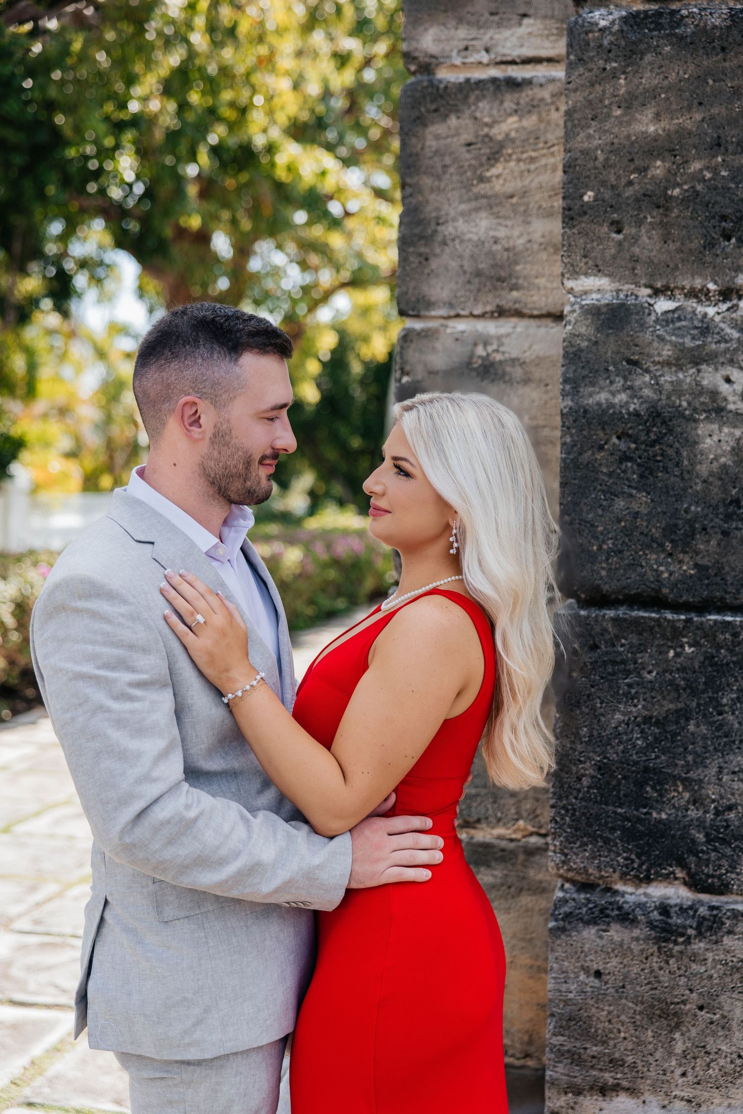 A couple embraces against a stone wall, with the woman wearing a red dress and the man in a light gray suit.