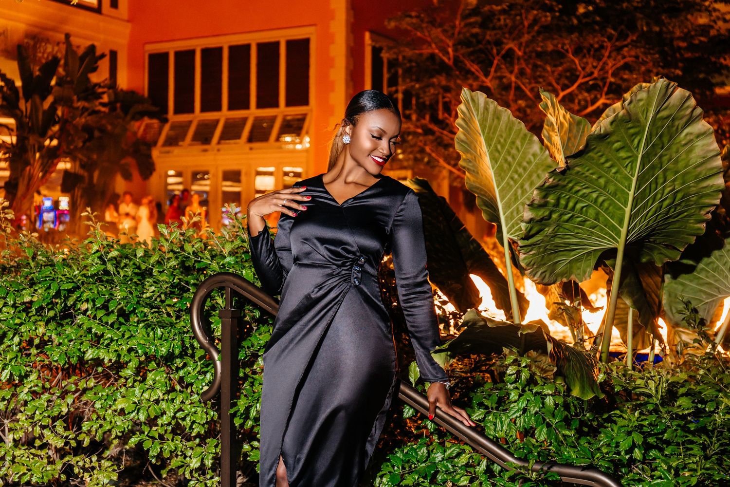 Woman in black dress poses near tropical plants with warm evening lighting.