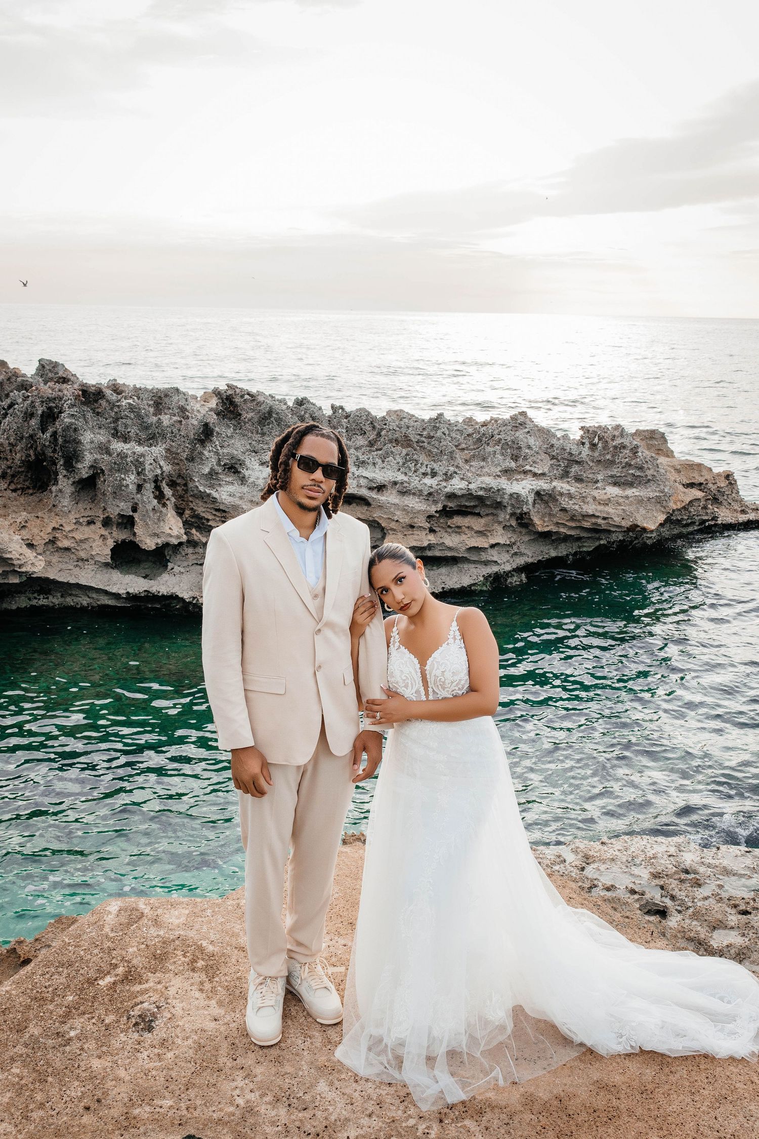 A wedding couple poses on rocky cliffs overlooking turquoise ocean waters in a scenic coastal setting.