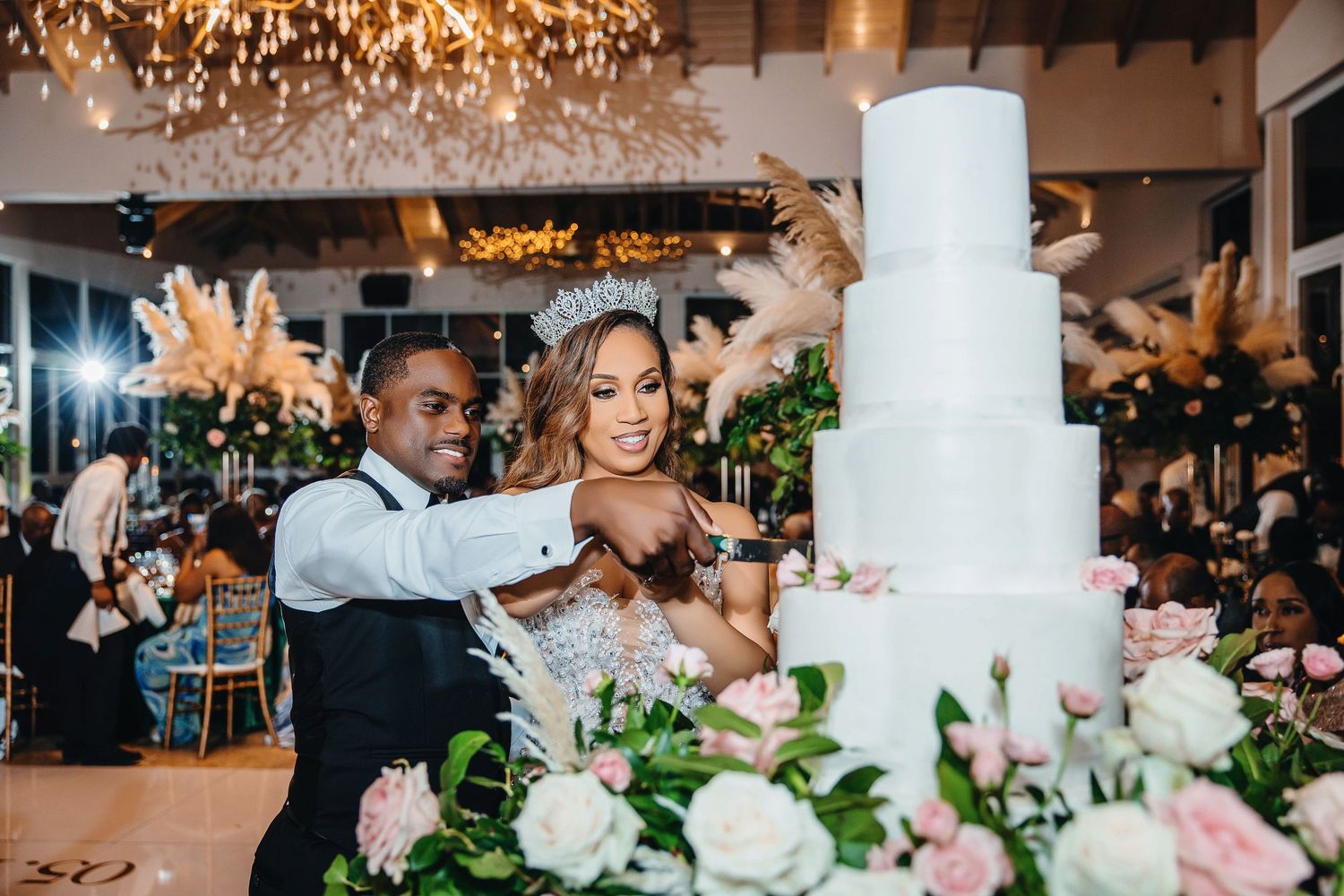 Newlyweds cut their elegant white wedding cake under crystal chandelier at reception.