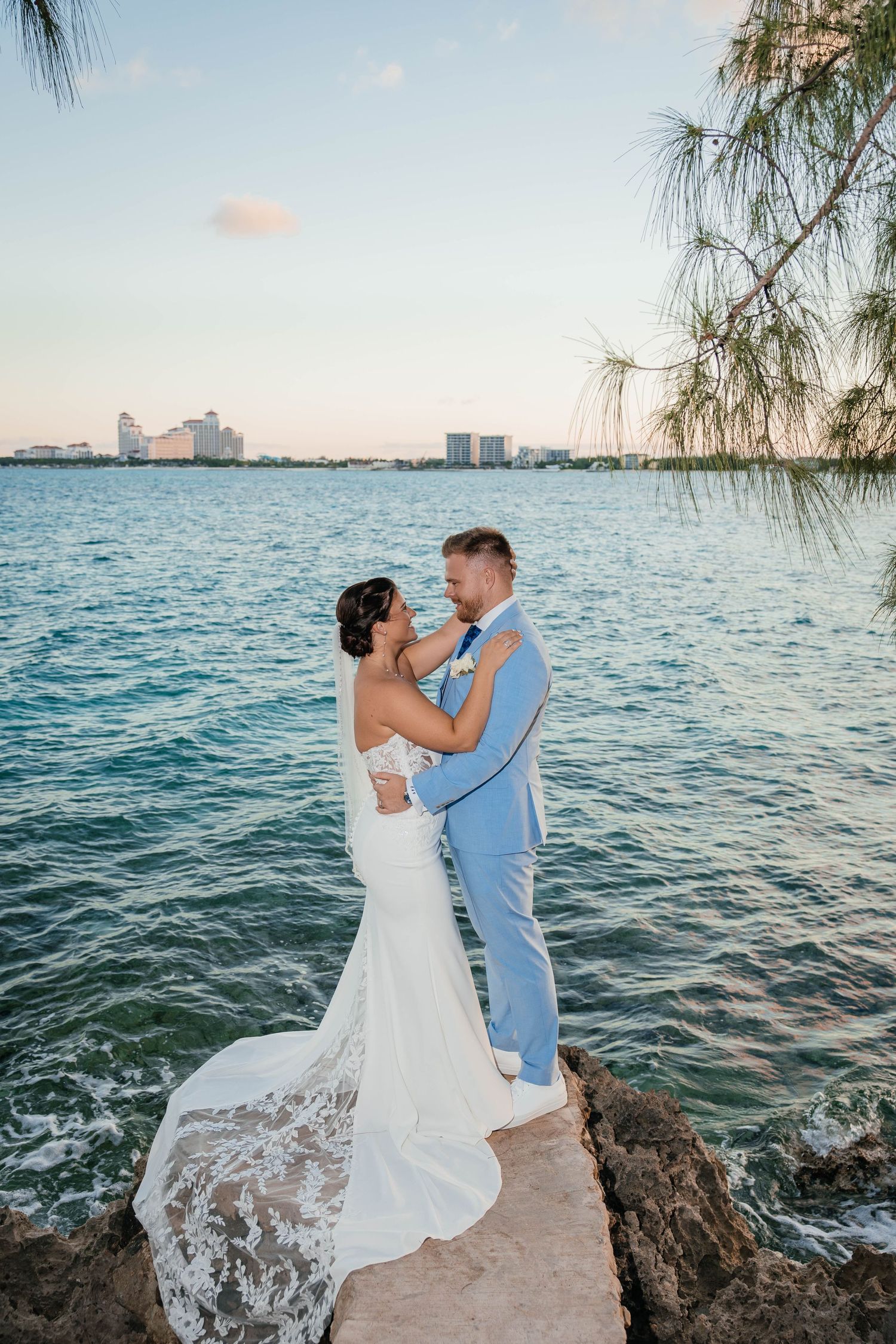 Newlyweds embrace on rocky oceanfront with blue waters and city skyline in the distance.