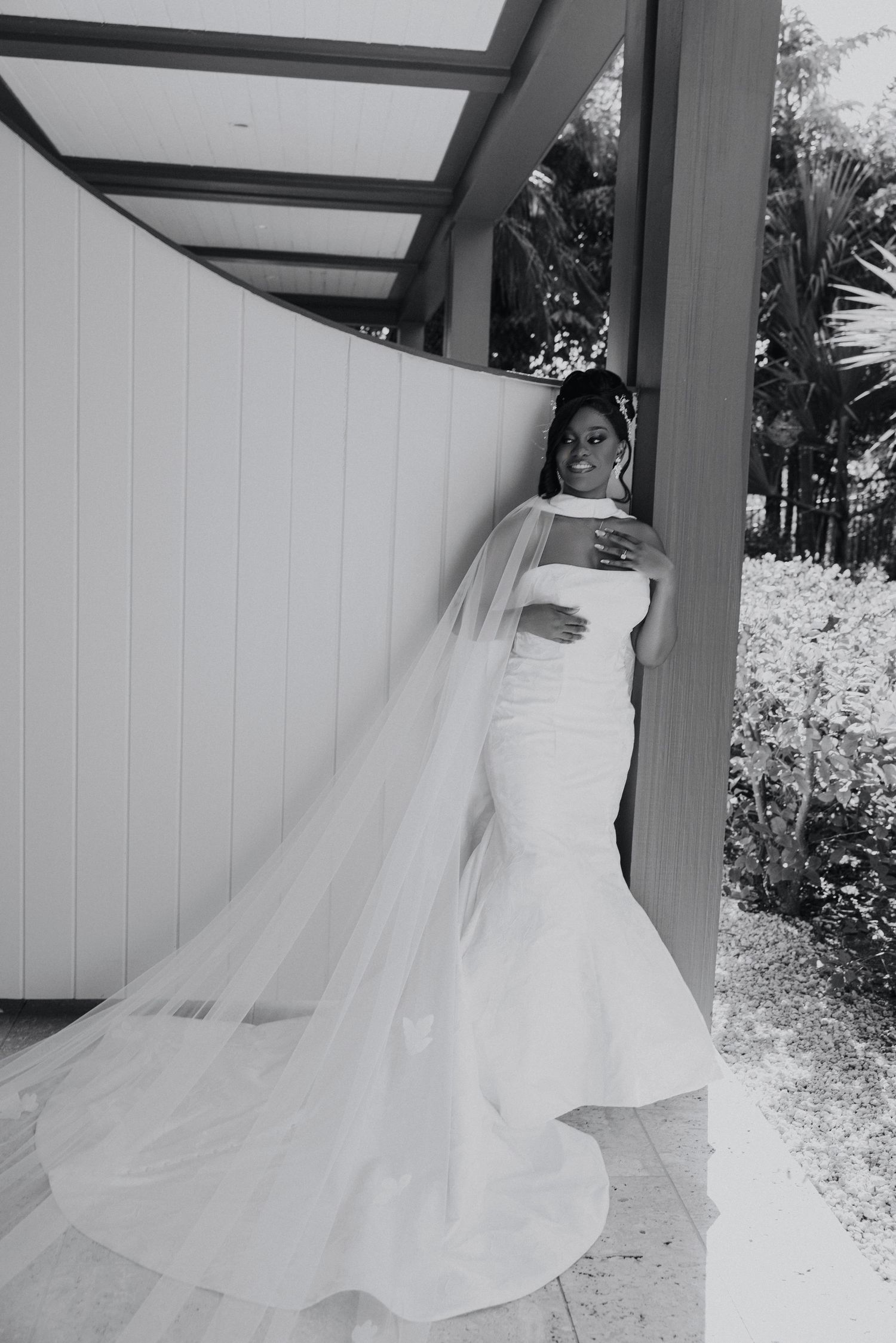 Black and white photo of a bride in a mermaid gown with a long veil, smiling outdoors near a white wall.