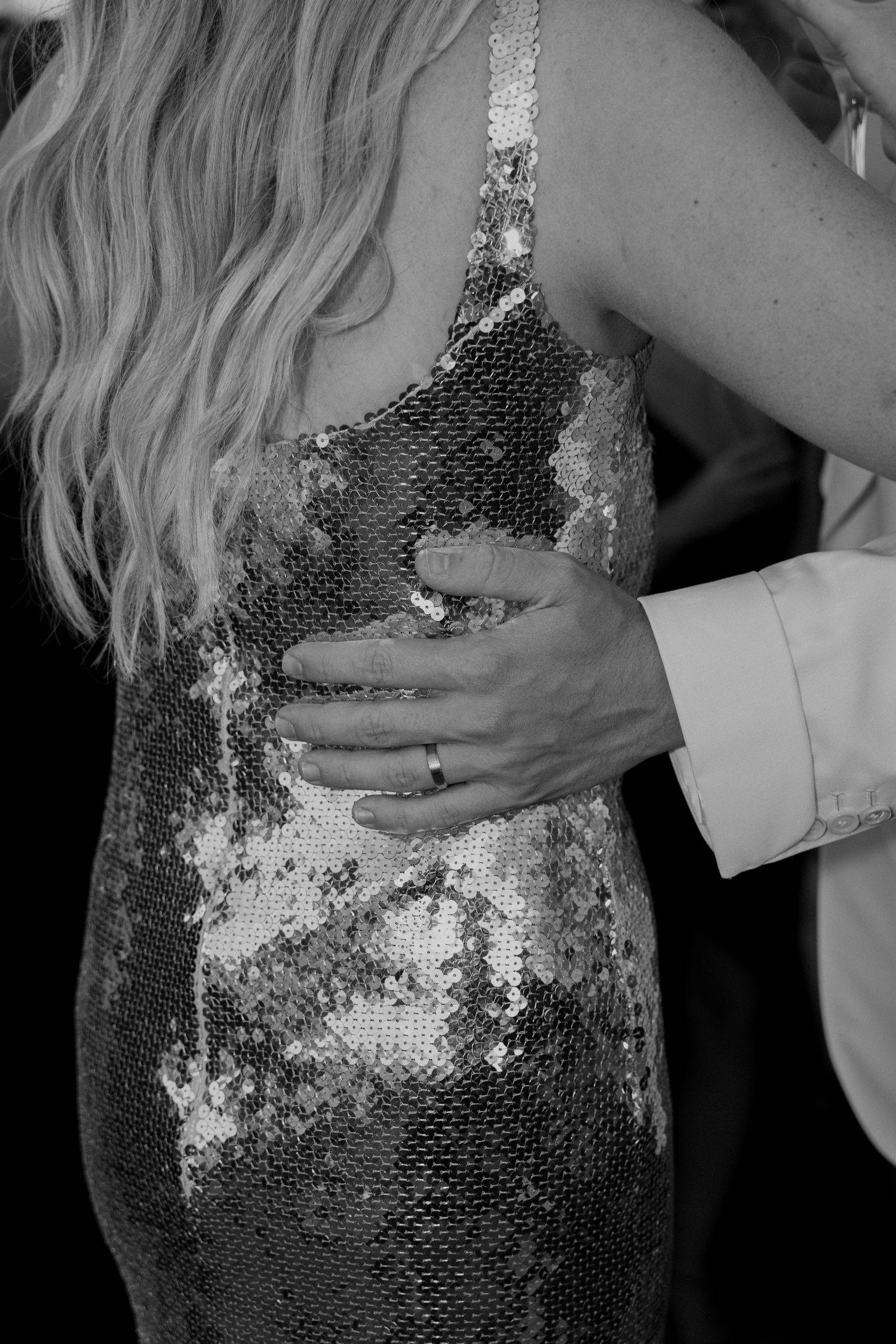 Black and white close-up of a man's hand with a wedding ring resting on the back of a woman in a sparkling sequin dress with long wavy hair.