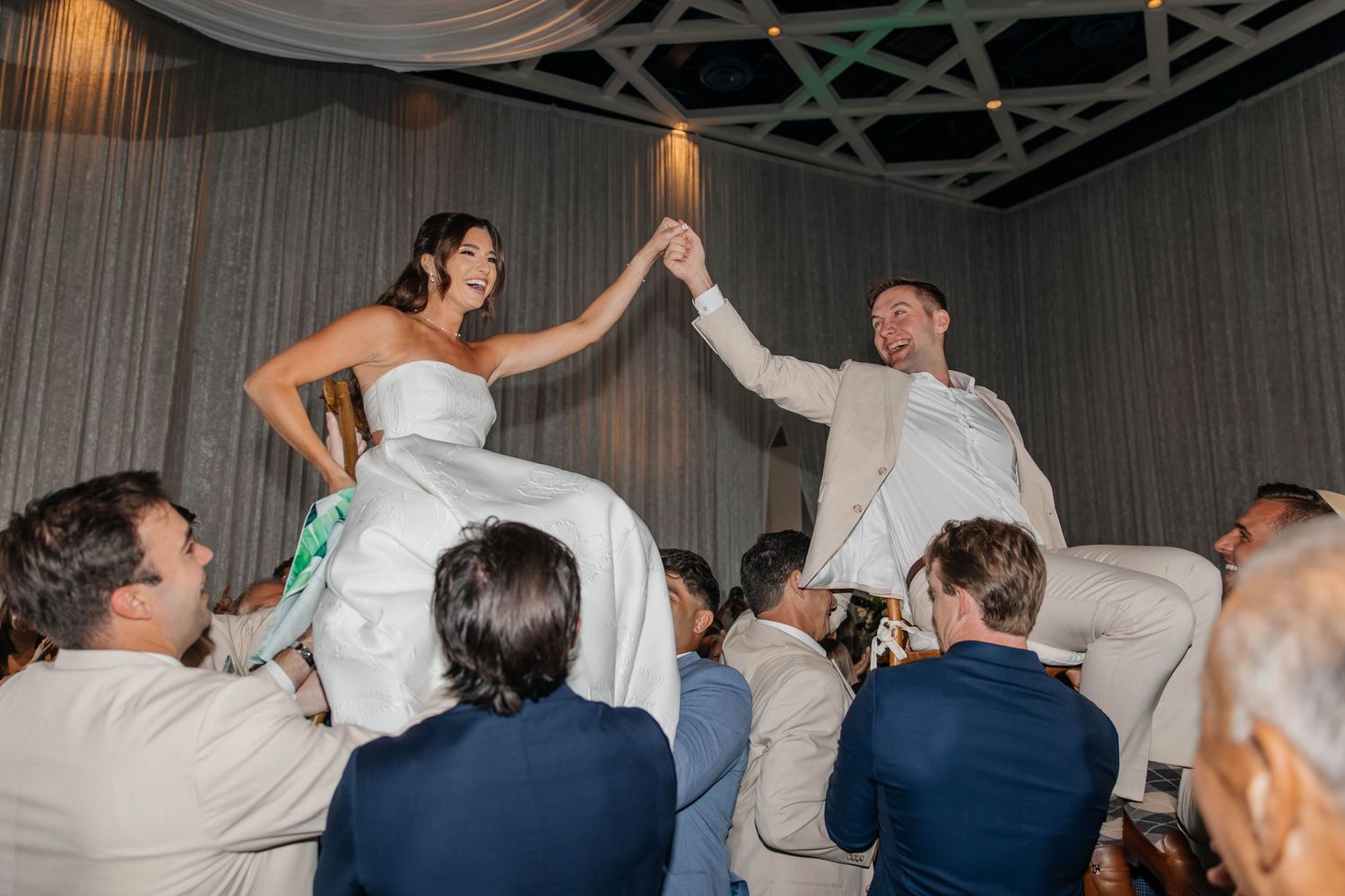 Bride and groom lifted on chairs during traditional Jewish Hora dance at elegant wedding reception.