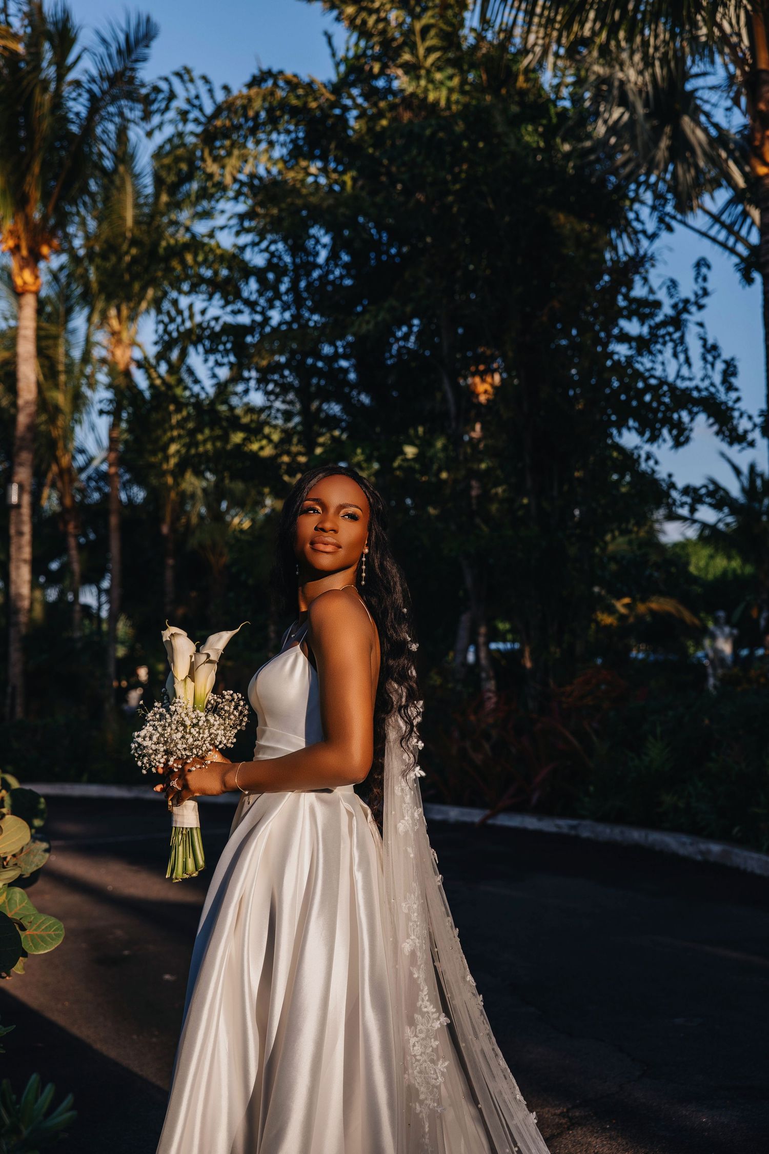 Elegant Black bride in a white satin gown holding a bouquet, posing among palm trees at golden hour.