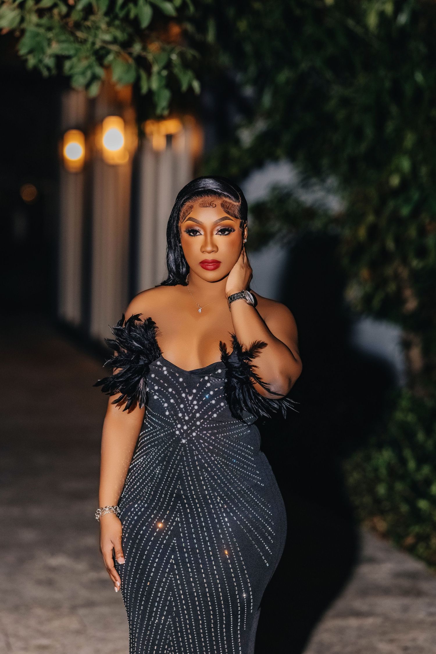 Glamorous evening portrait of a model in black feathered gown against dark outdoor setting.