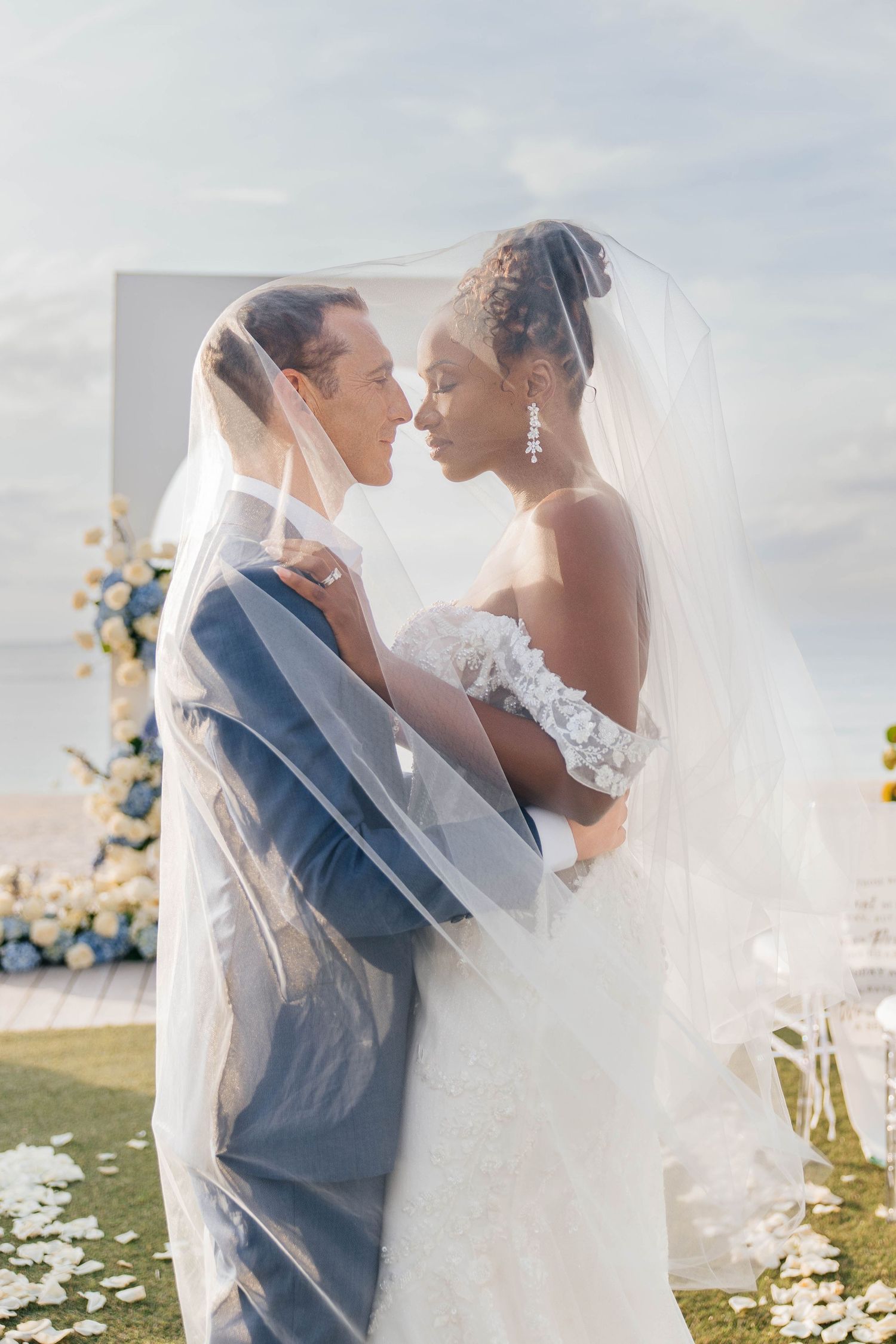 A romantic wedding couple embraces outdoors in soft sunlight with flowing white veil and blue suit.