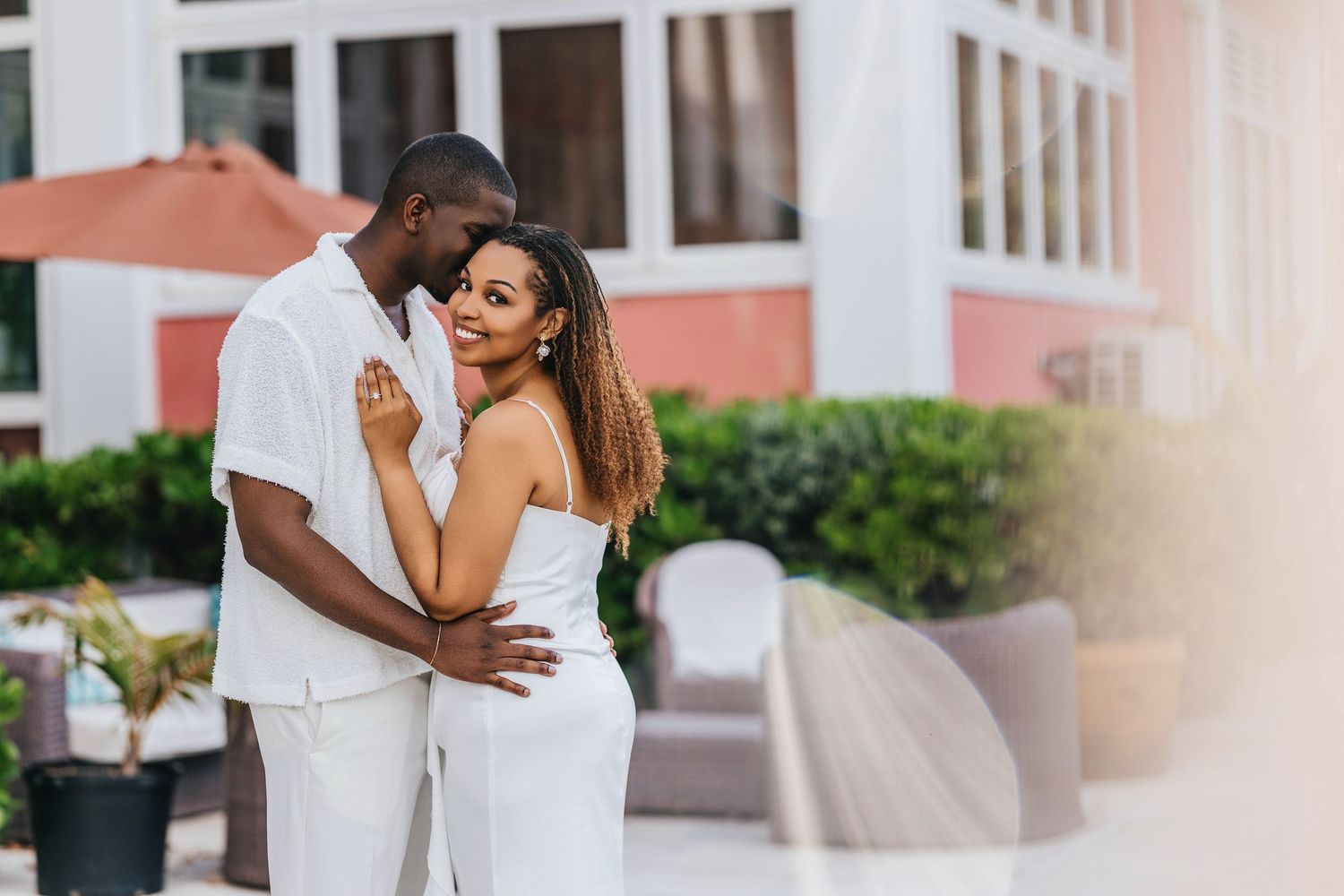 A romantic couple in white outfits embrace while standing outside a pink building on a sunny day.