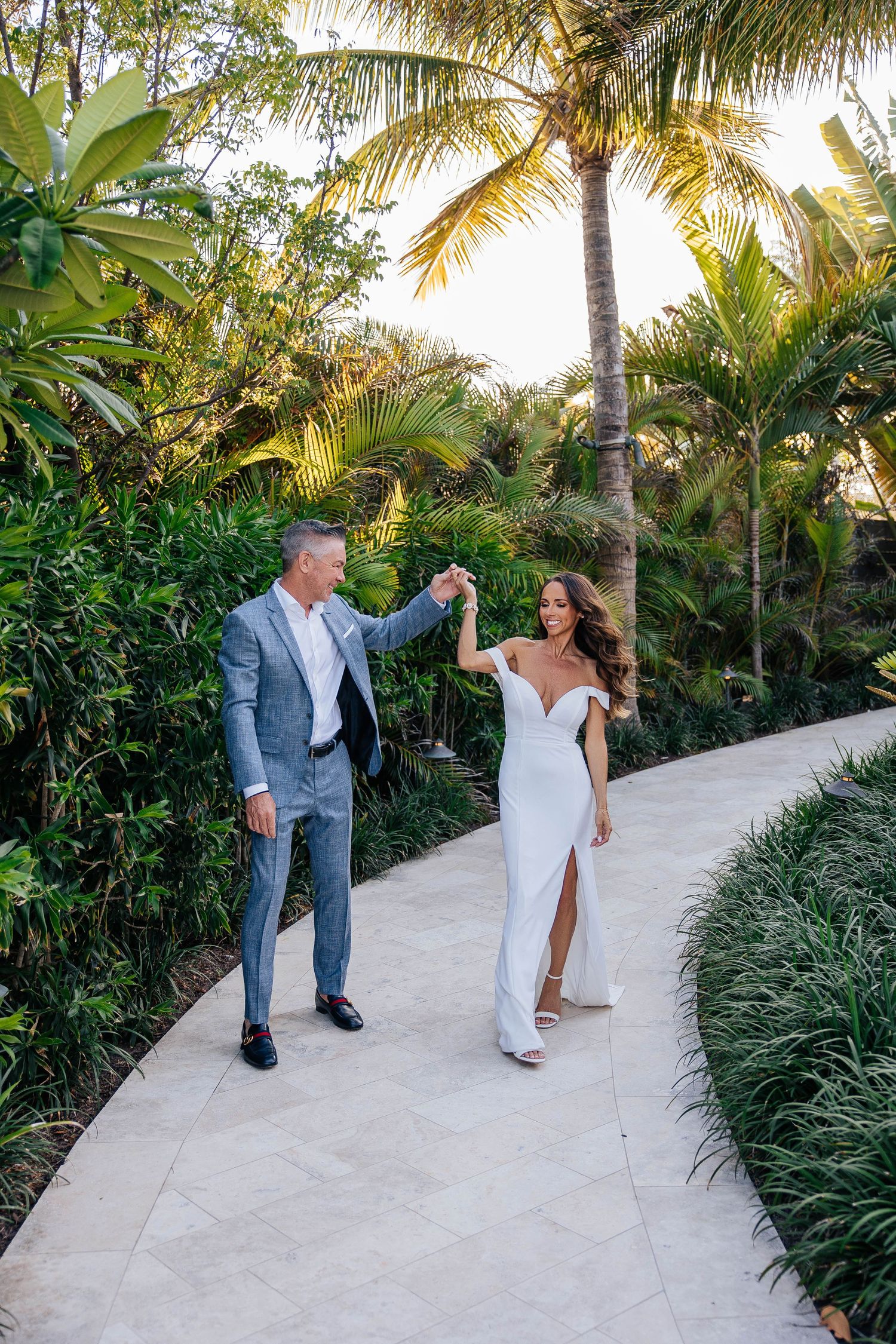Newlyweds dancing together on white path surrounded by tropical palms and greenery.