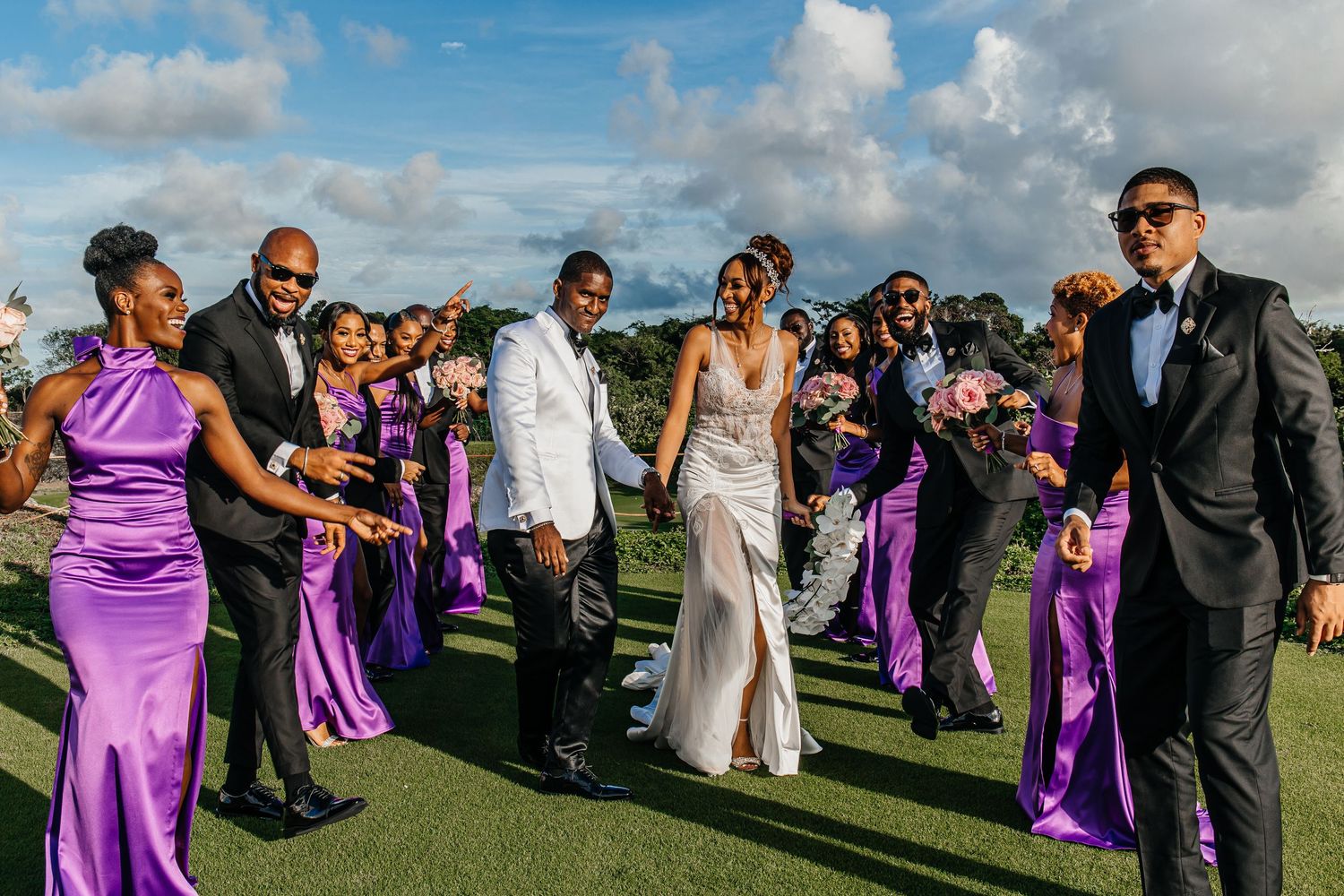 Wedding party dances and celebrates outdoors with bridesmaids wearing bright purple satin dresses.