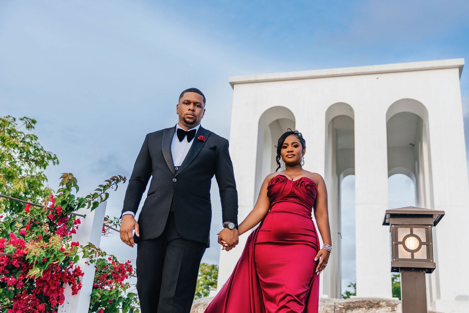 Couple in formal wear standing beneath white arched architecture with red flowers.
