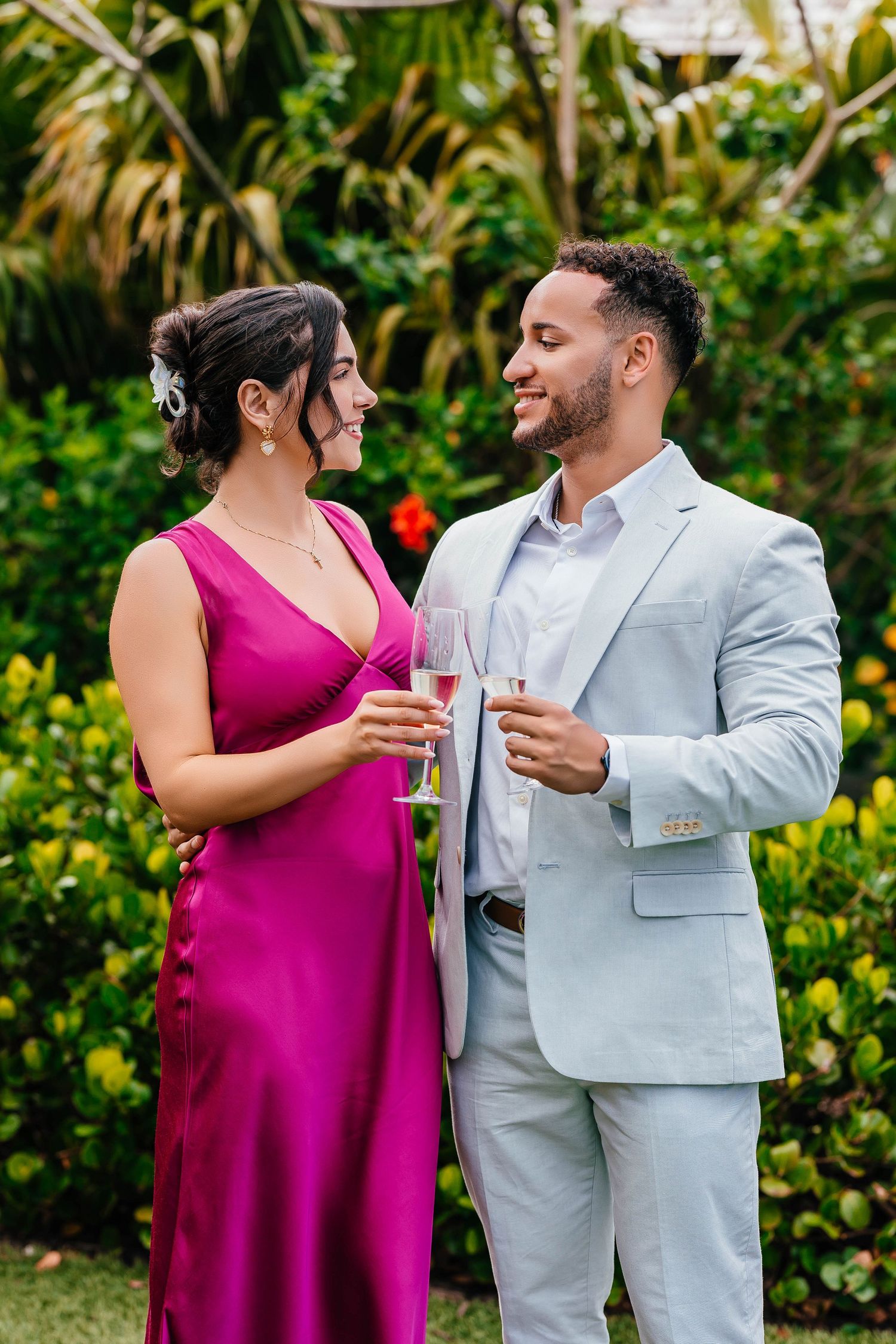 A couple shares drinks and laughter in formal attire against a lush tropical garden backdrop.