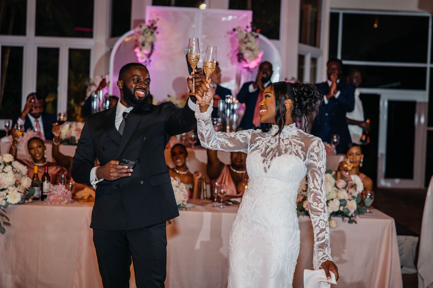 Bride in lace gown and groom in black tuxedo raising champagne glasses in toast at elegant wedding reception.