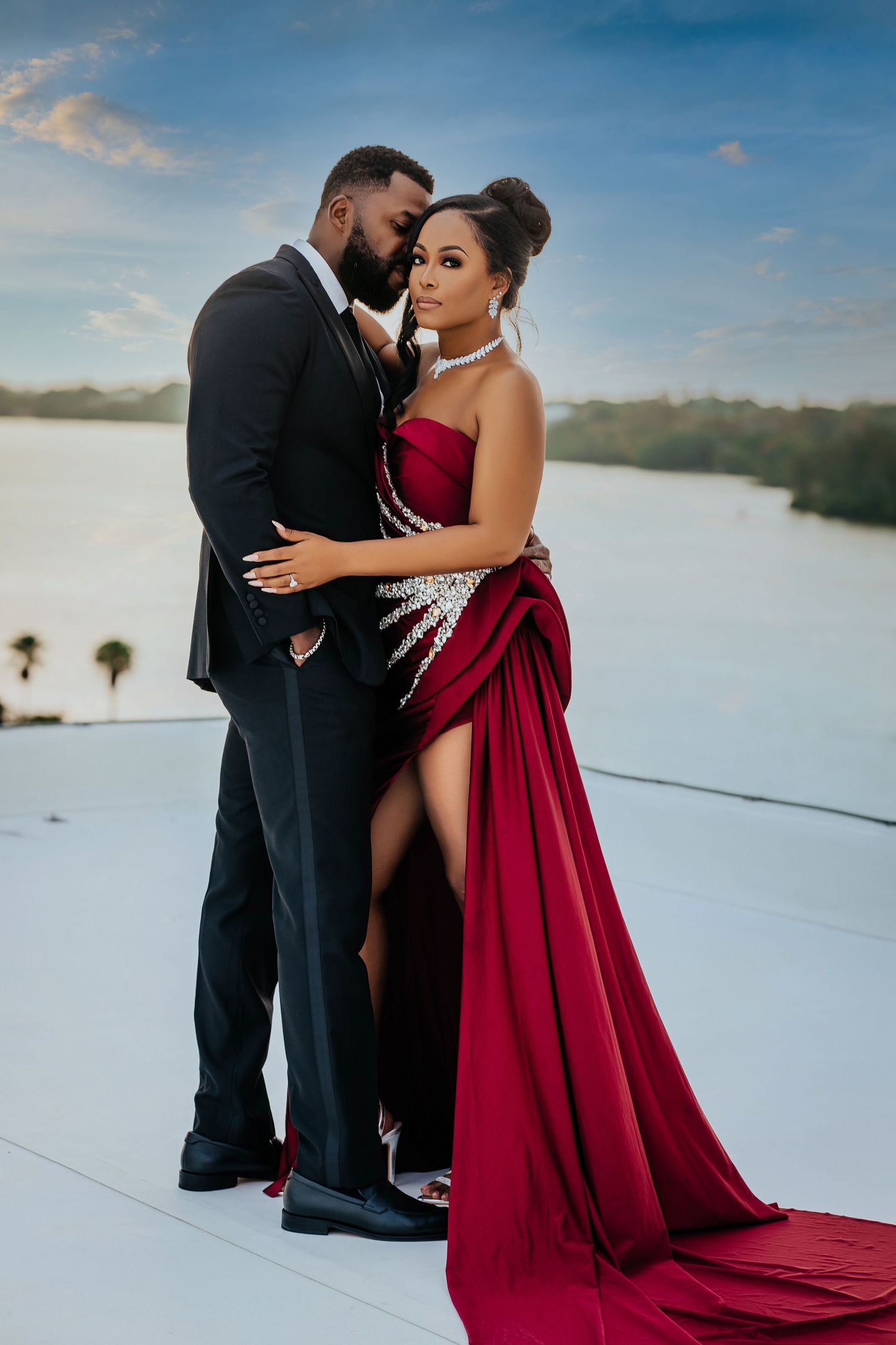 Romantic couple in formal attire posing against waterfront backdrop during sunset.