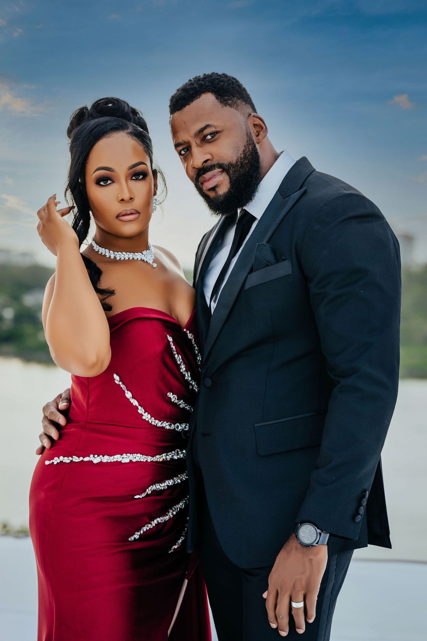 Glamorous couple poses against sunset sky in red sparkle gown and black tuxedo.