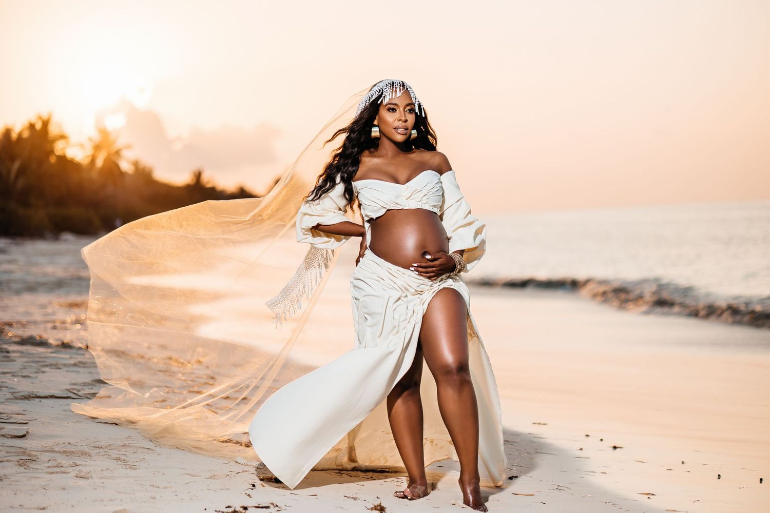 Dramatic maternity photo with flowing white dress during beach sunset.