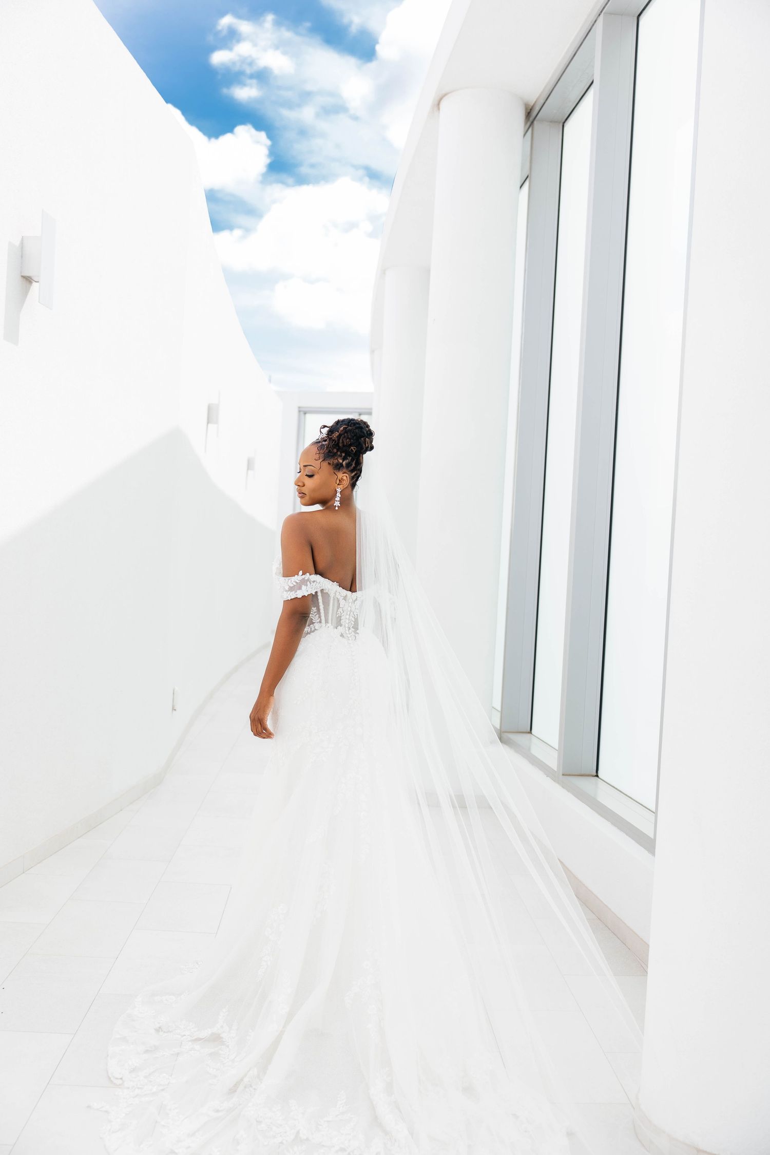 A person in a white dress walks down a narrow white hallway with blue sky visible through windows.