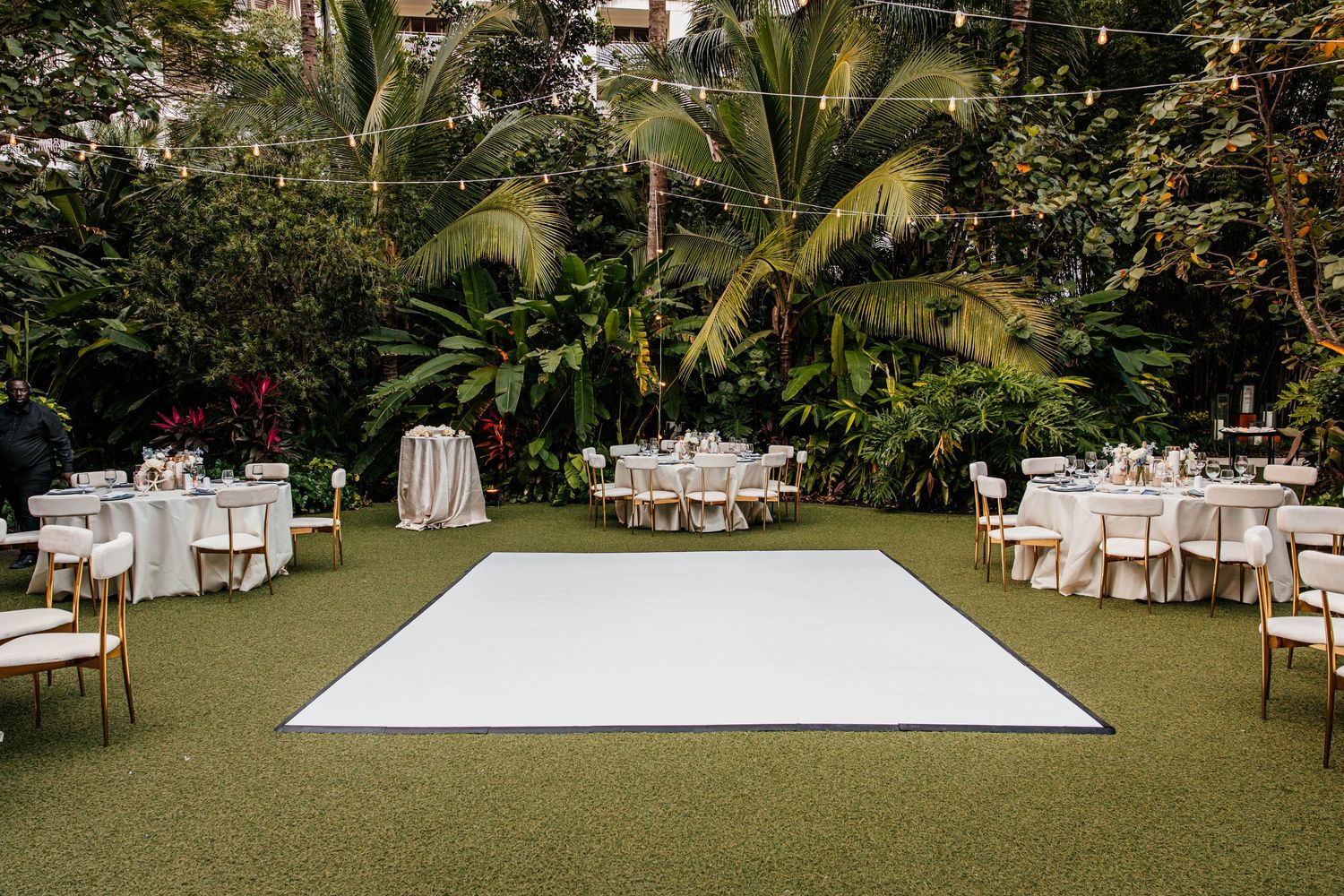 Outdoor wedding reception with white dance floor, round tables, gold chairs, and lush tropical greenery backdrop.