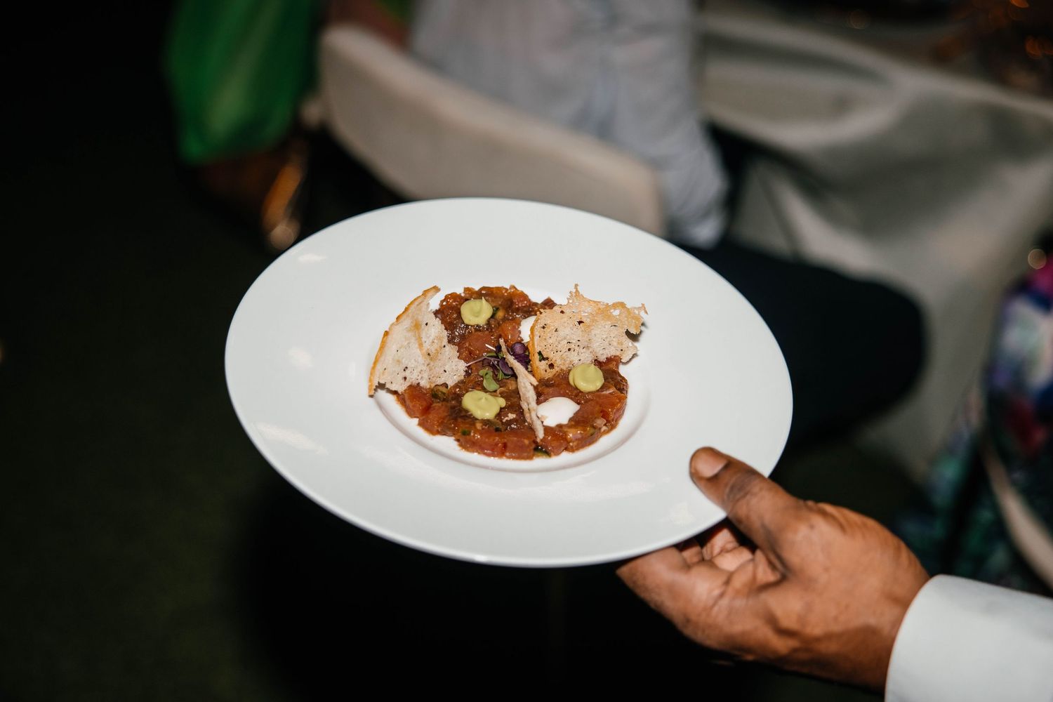 A waiter holds a white plate with an elegant appetizer featuring tomato sauce, bread crisps, and garnishes.