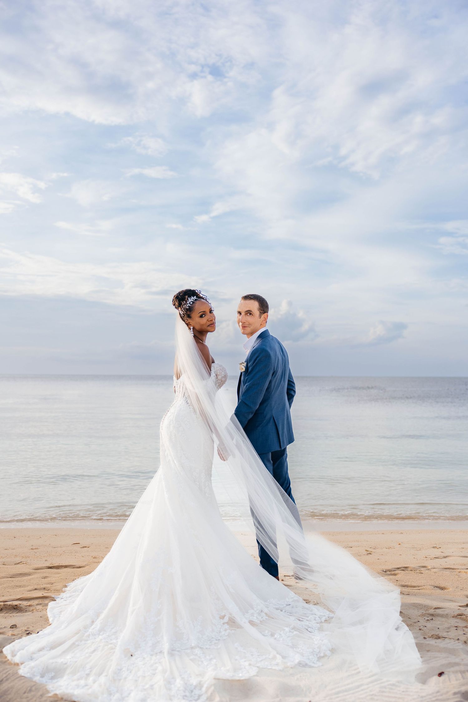 A newlywed couple poses for photographs on a beach with flowing wedding dress and veil against a serene ocean backdrop.