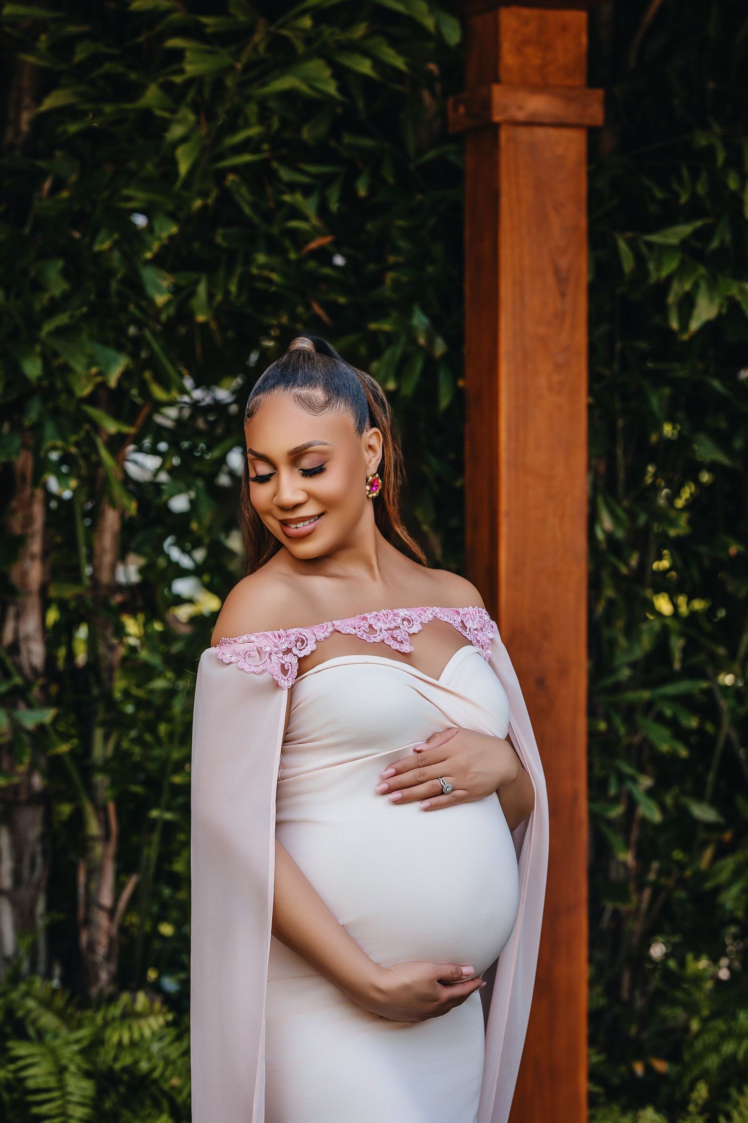 Elegant maternity portrait featuring a white off-shoulder gown with pink accents in a garden setting.