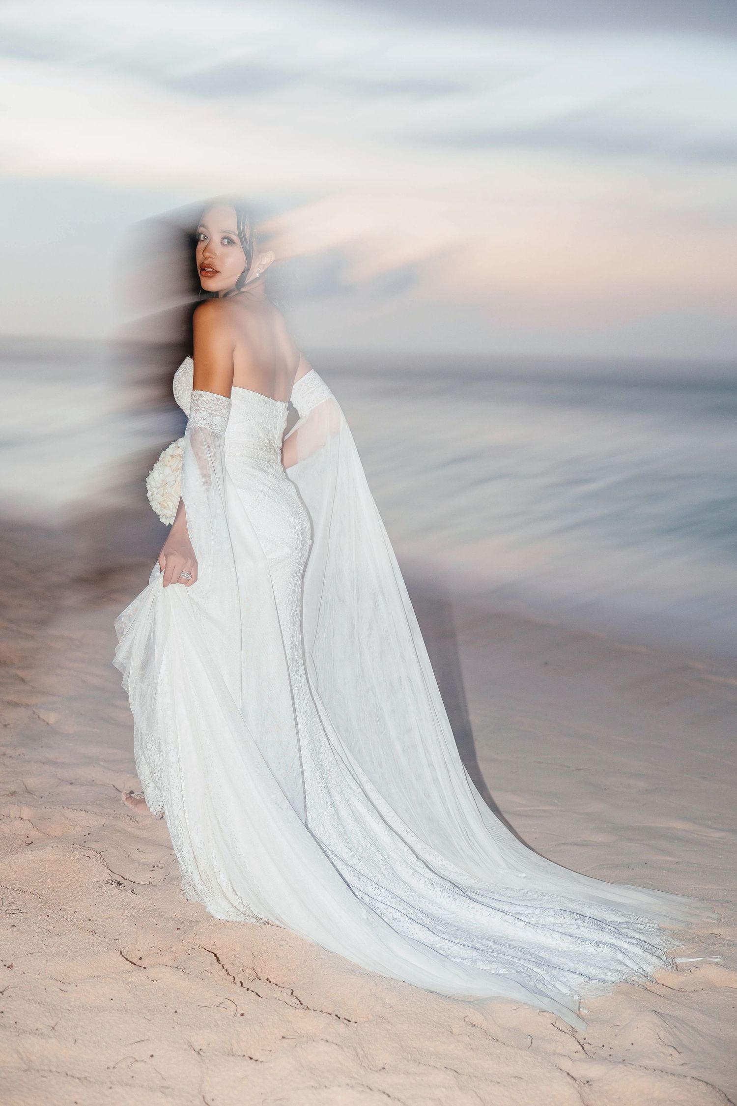 Bride in flowing white gown with long train stands on sandy beach at sunset, looking over her shoulder.