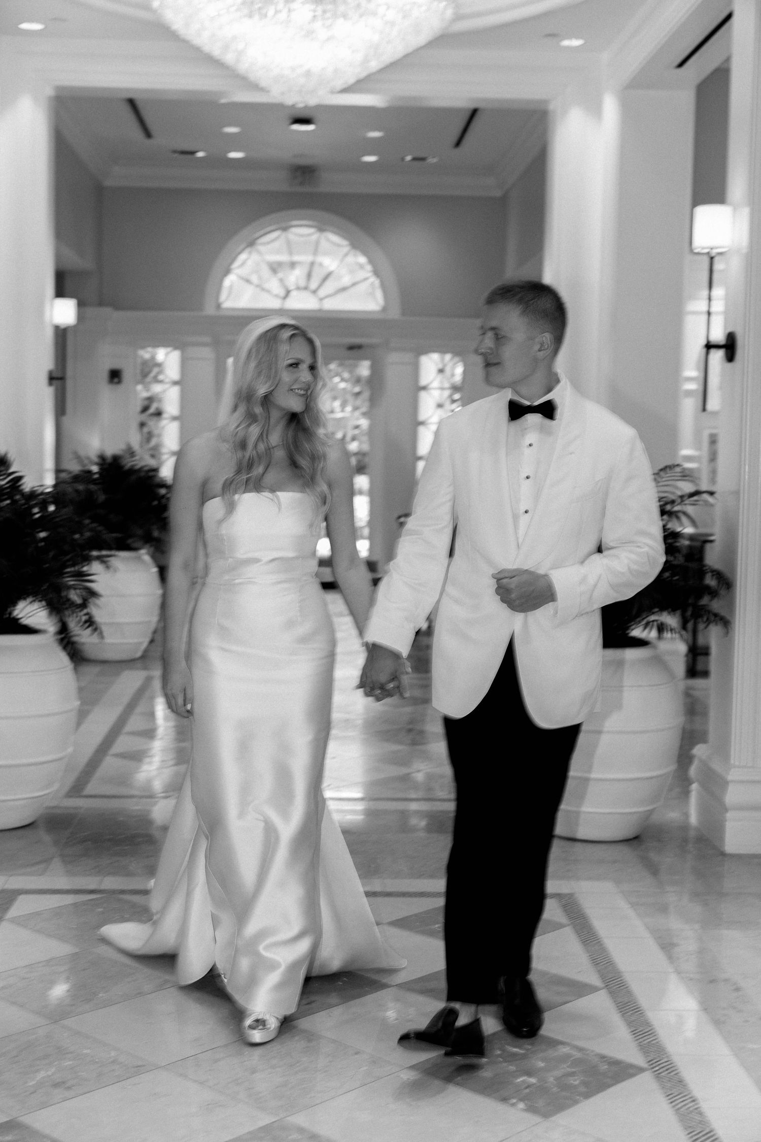 Black and white photo of a bride in a satin gown holding hands with a groom in a white dinner jacket and bow tie in an elegant hallway.