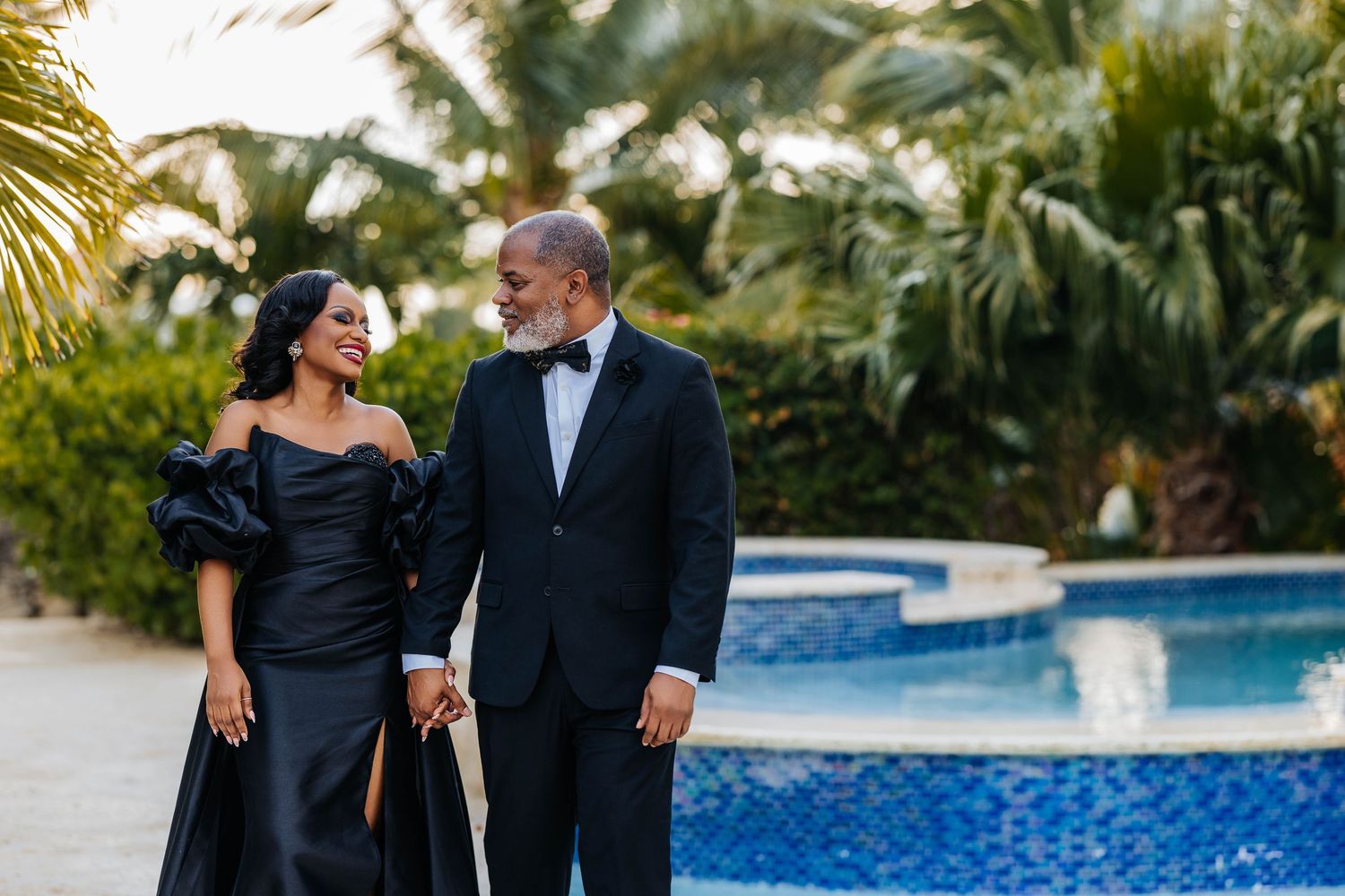 Couple in elegant black formal attire walking and posing together by a pool at sunset during a photoshoot.