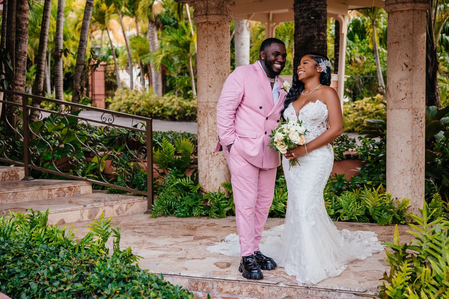 Romantic wedding portrait with pink suit and white lace gown in tropical garden setting.
