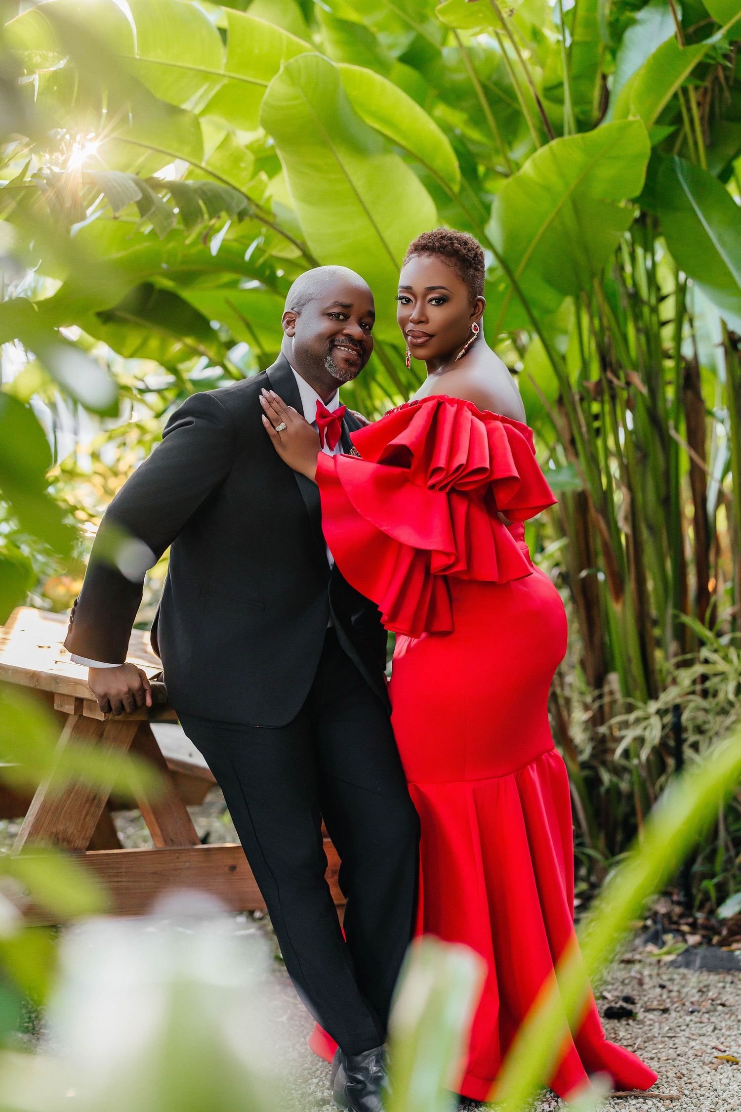 Couple in formal attire poses together among tropical foliage with dramatic red dress.